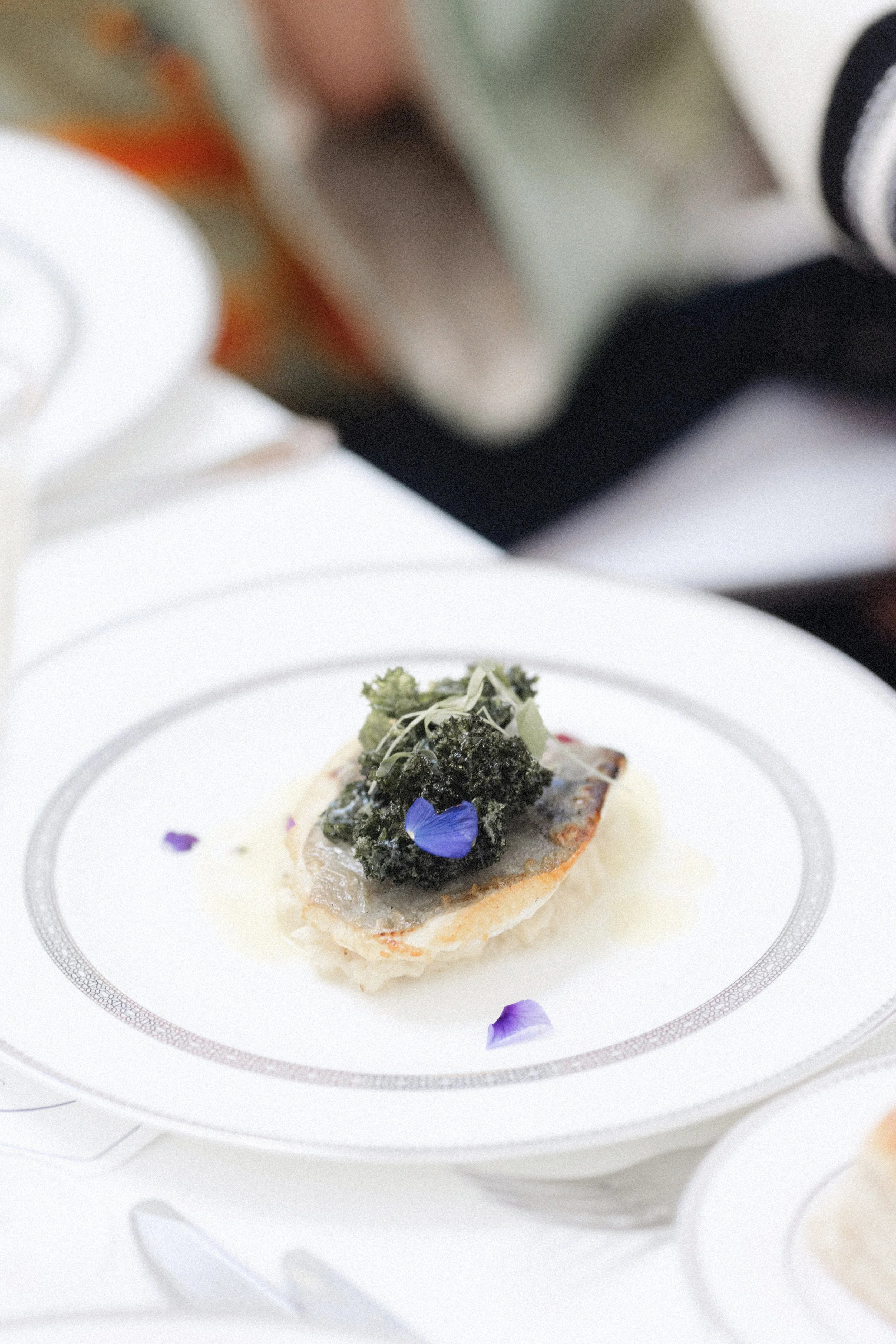 A gourmet appetizer with a small piece of bread topped with pesto, microgreens, edible flowers, and purple flower petals on a white plate.