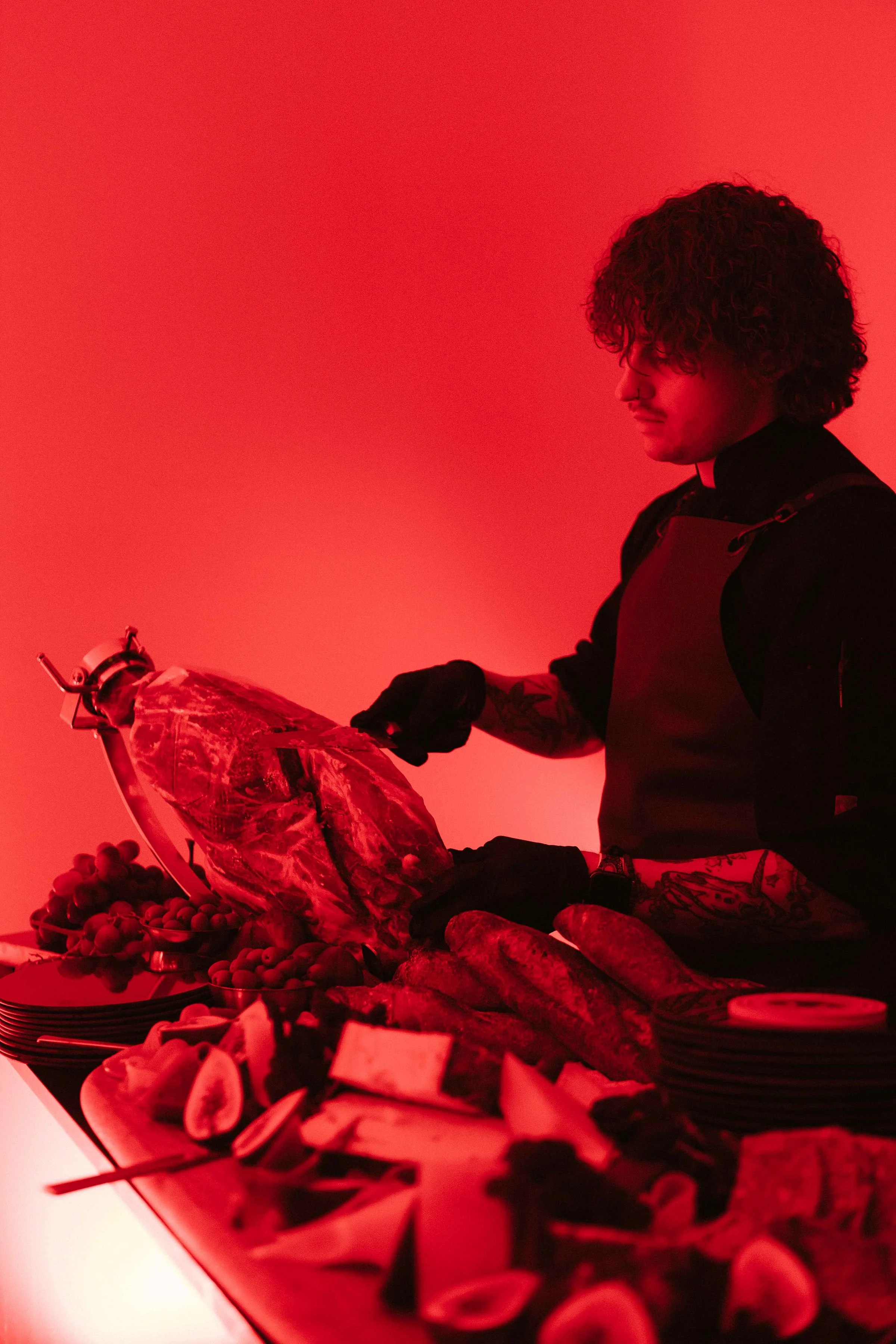 A person with curly hair and tattoos cutting a large piece of meat at a table full of food, with a red lighting background.
