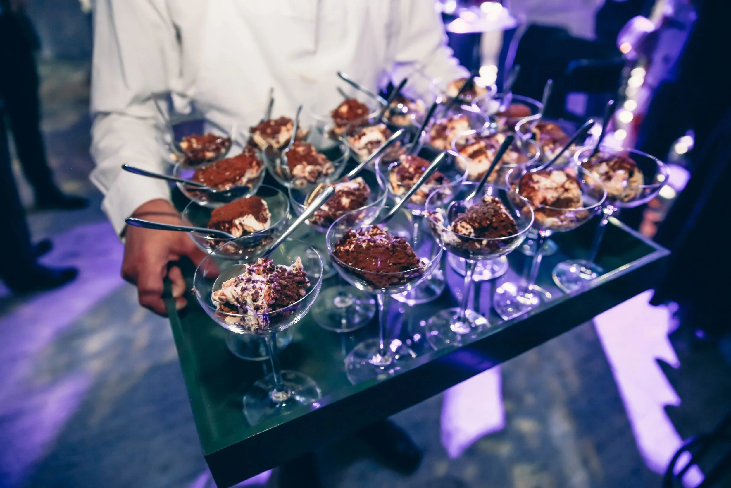 A person holding a tray of small dessert glasses filled with chocolate and cream treats at a party or event.