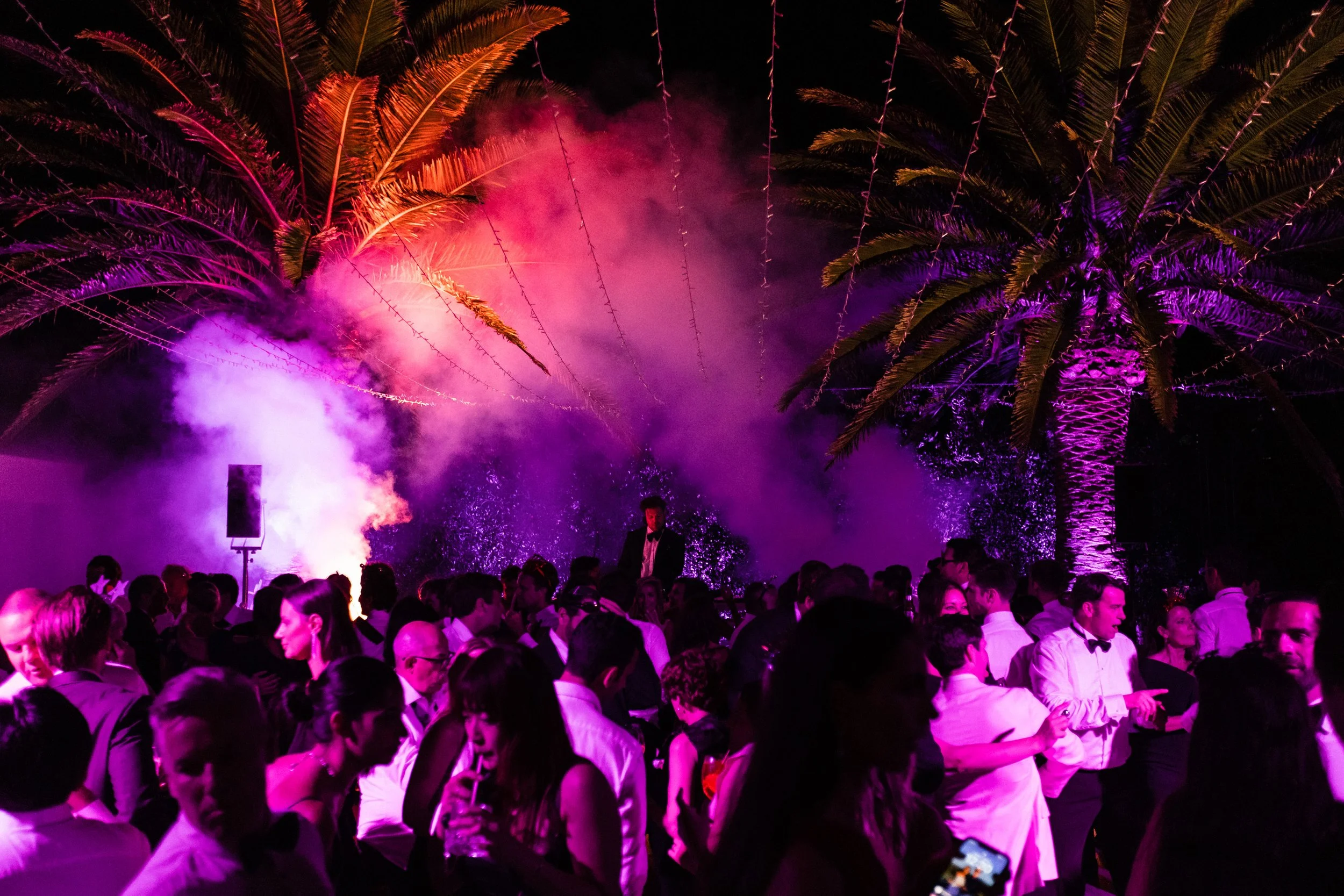 Wedding guests dancing under palm trees lit in pink and purple haze at a New Year's Eve celebration in Sorrento