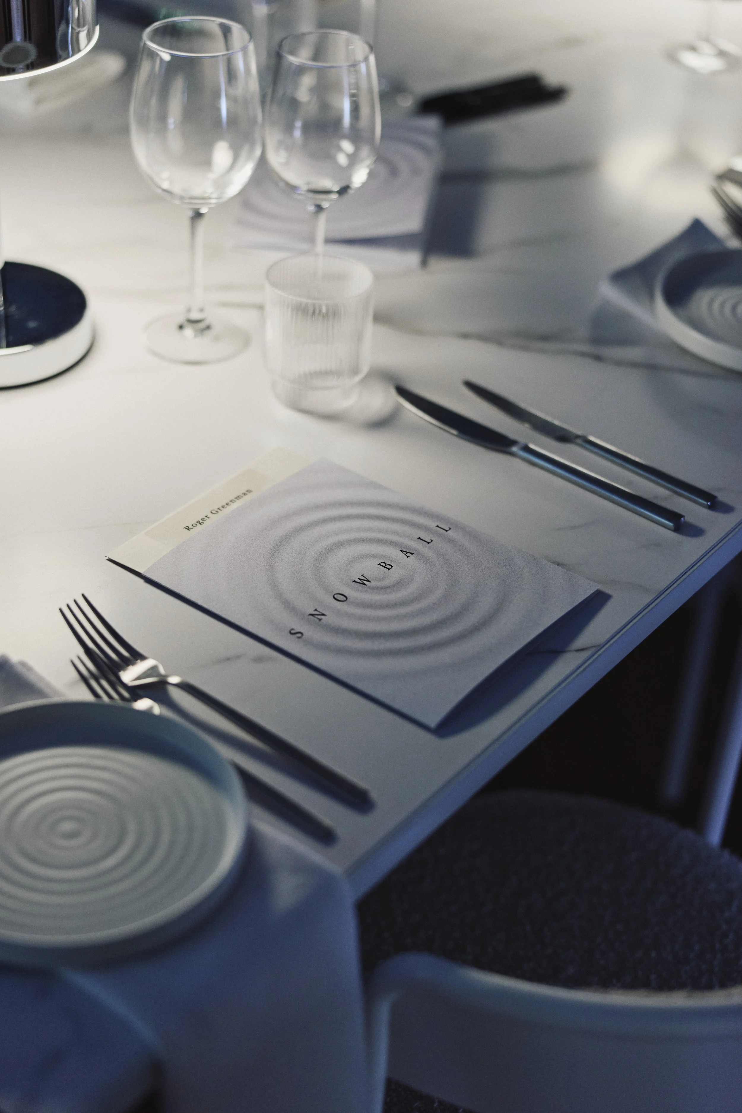 A dining table set with two empty wine glasses, a clear drinking glass, a menu, and silverware, with a white tablecloth.