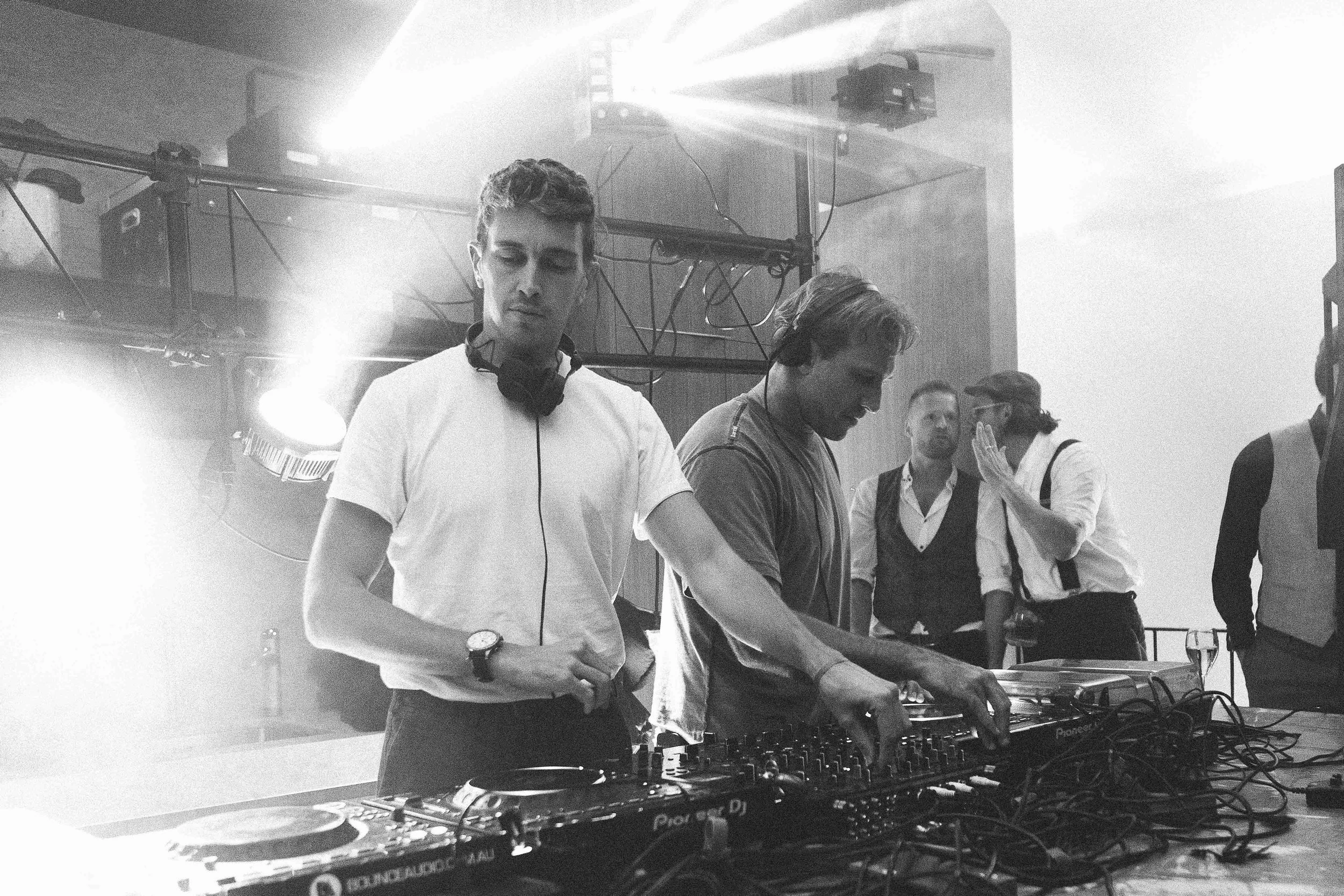 A group of young people, including a DJ, setting up DJ equipment at an indoor event or party with bright lighting and a modern industrial interior.