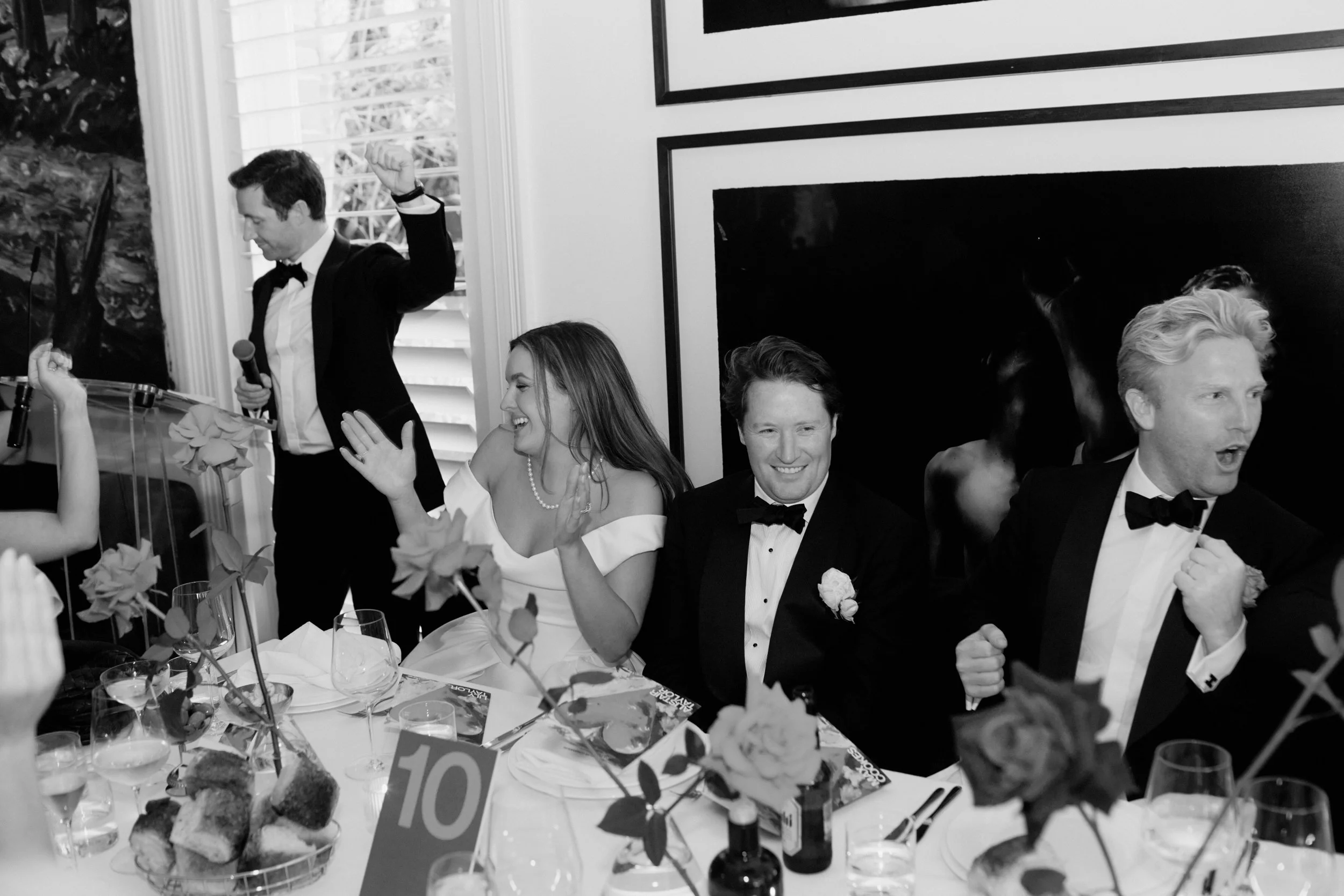 A group of formally dressed people at a celebration or wedding, some are smiling and talking, seated around a table with flowers, drinks, and a cake, with a man standing and speaking into a microphone.