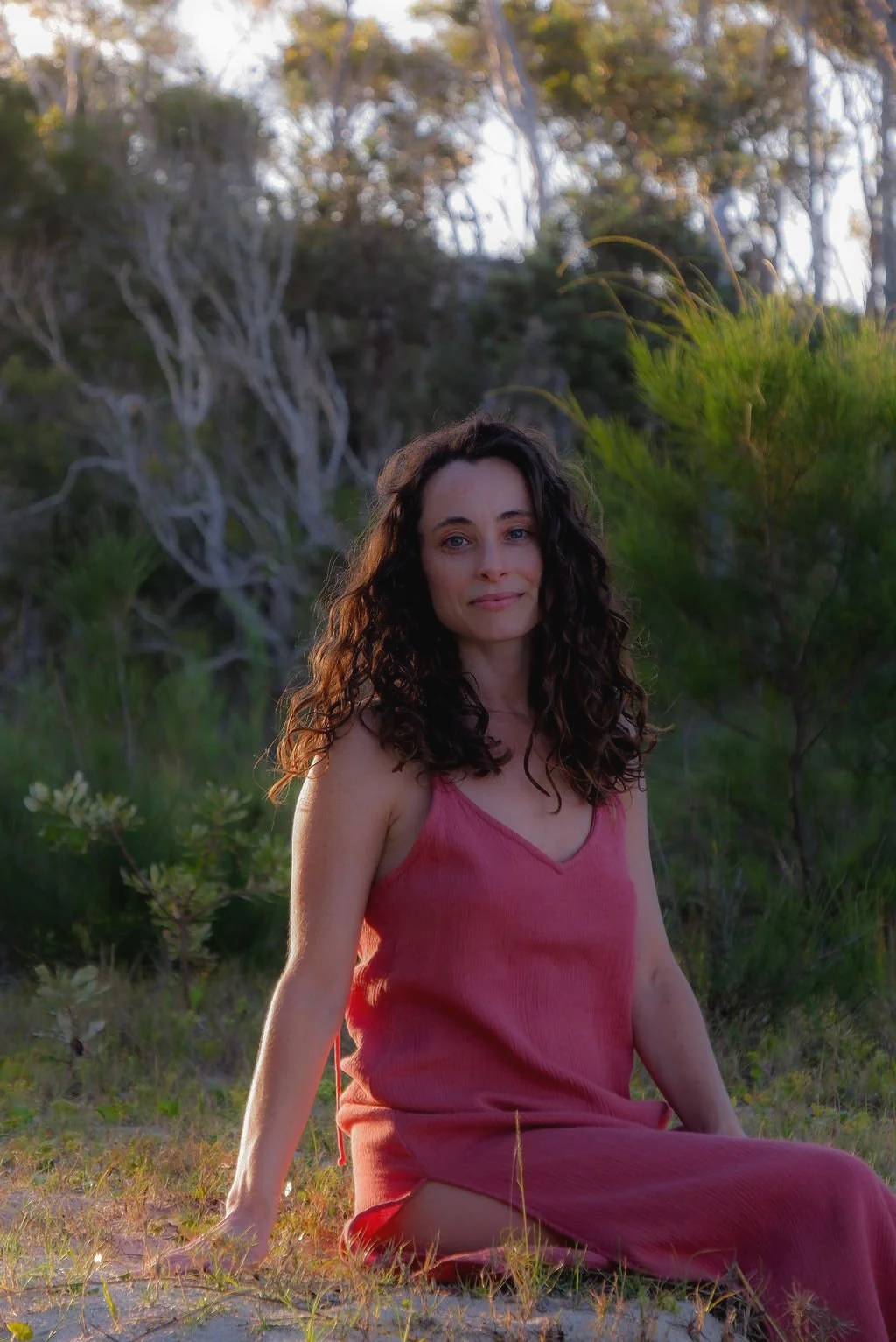A woman with long curly dark hair in a pink dress sitting outdoors in a natural setting with green trees and bushes.