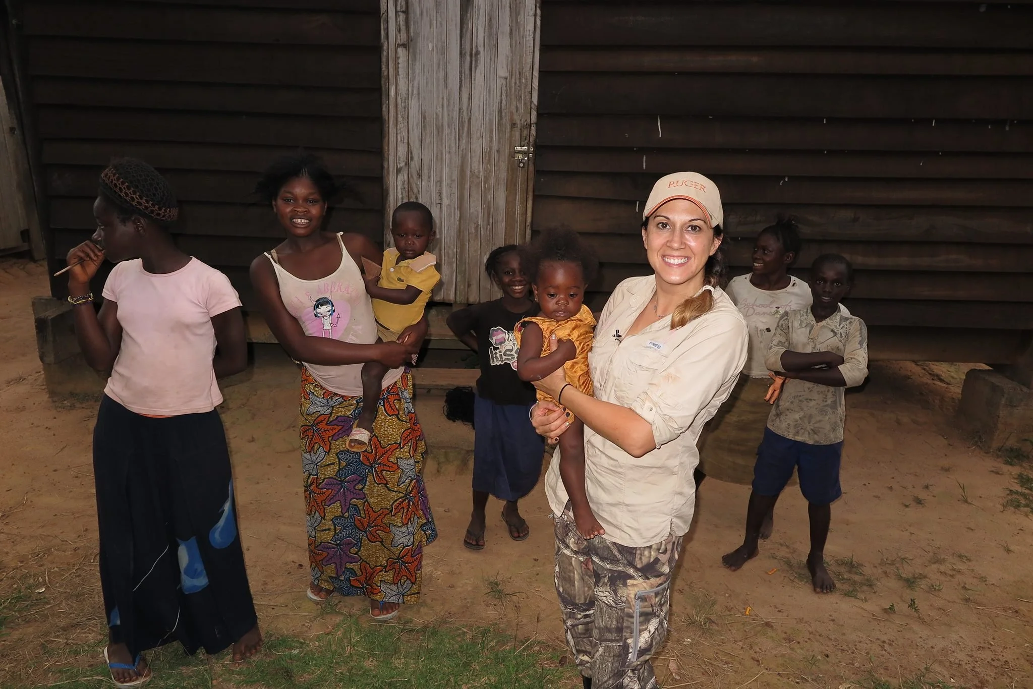 A woman holding a young girl smiles at the camera, surrounded by six children standing in front of a wooden building in a rural area.