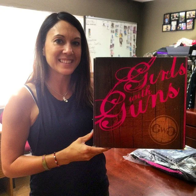 A woman with long dark hair wearing a sleeveless black top, smiling and holding a box labeled 'Girls with Guns' in a room with a whiteboard and photo collage in the background.