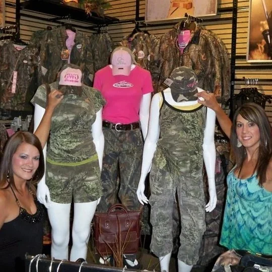 Display of camouflage clothing, hats, and a pink T-shirt in a store, with two women smiling and pointing at the items.