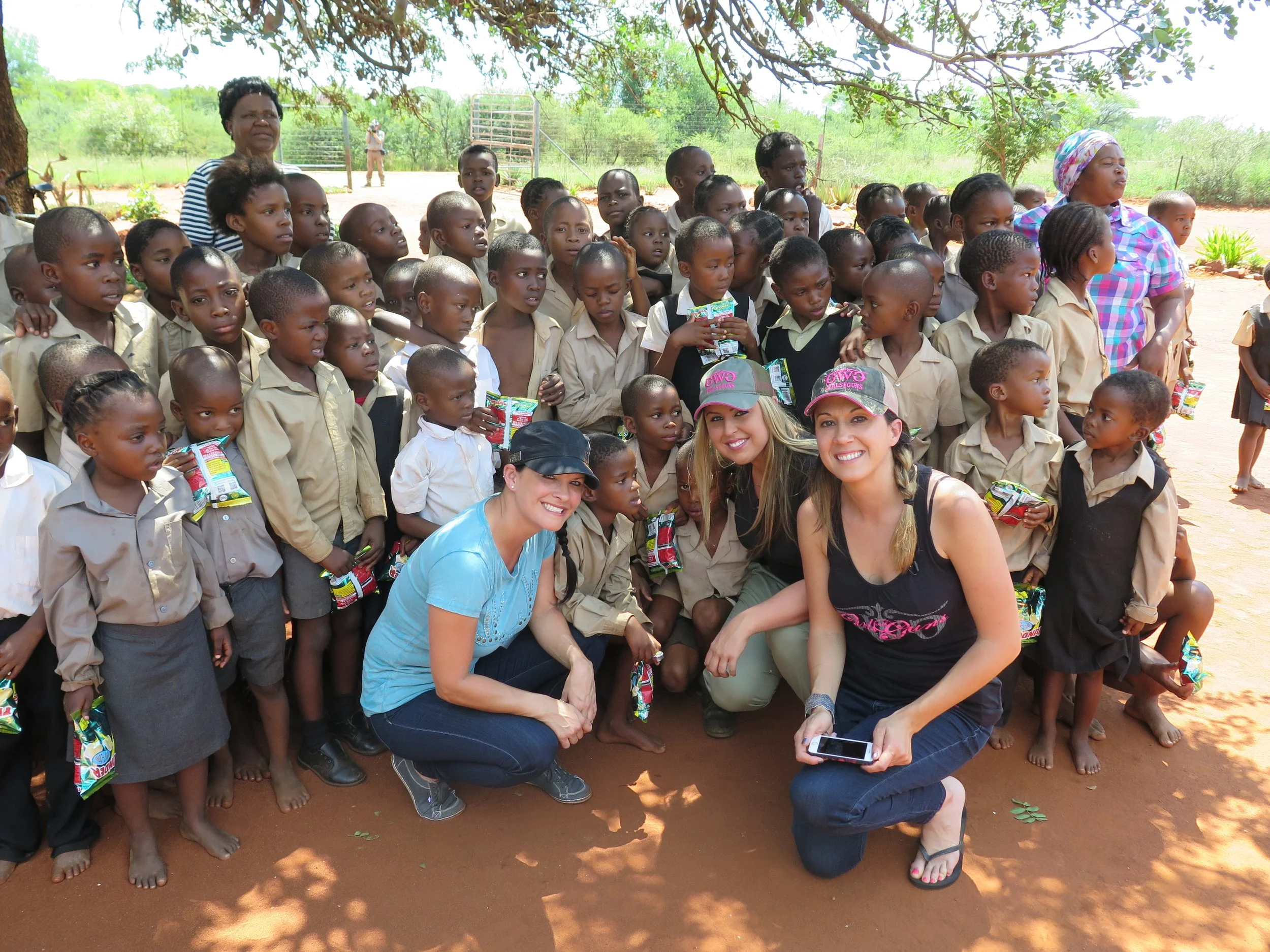 Group of children in school uniforms with three women, two crouching and one standing, under a tree outdoors in a rural setting.