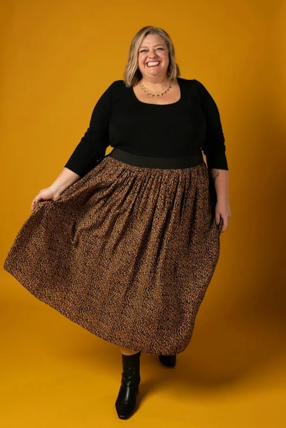 A smiling woman with blonde hair wearing a black top, a leopard print skirt, and black boots, standing against a yellow background.