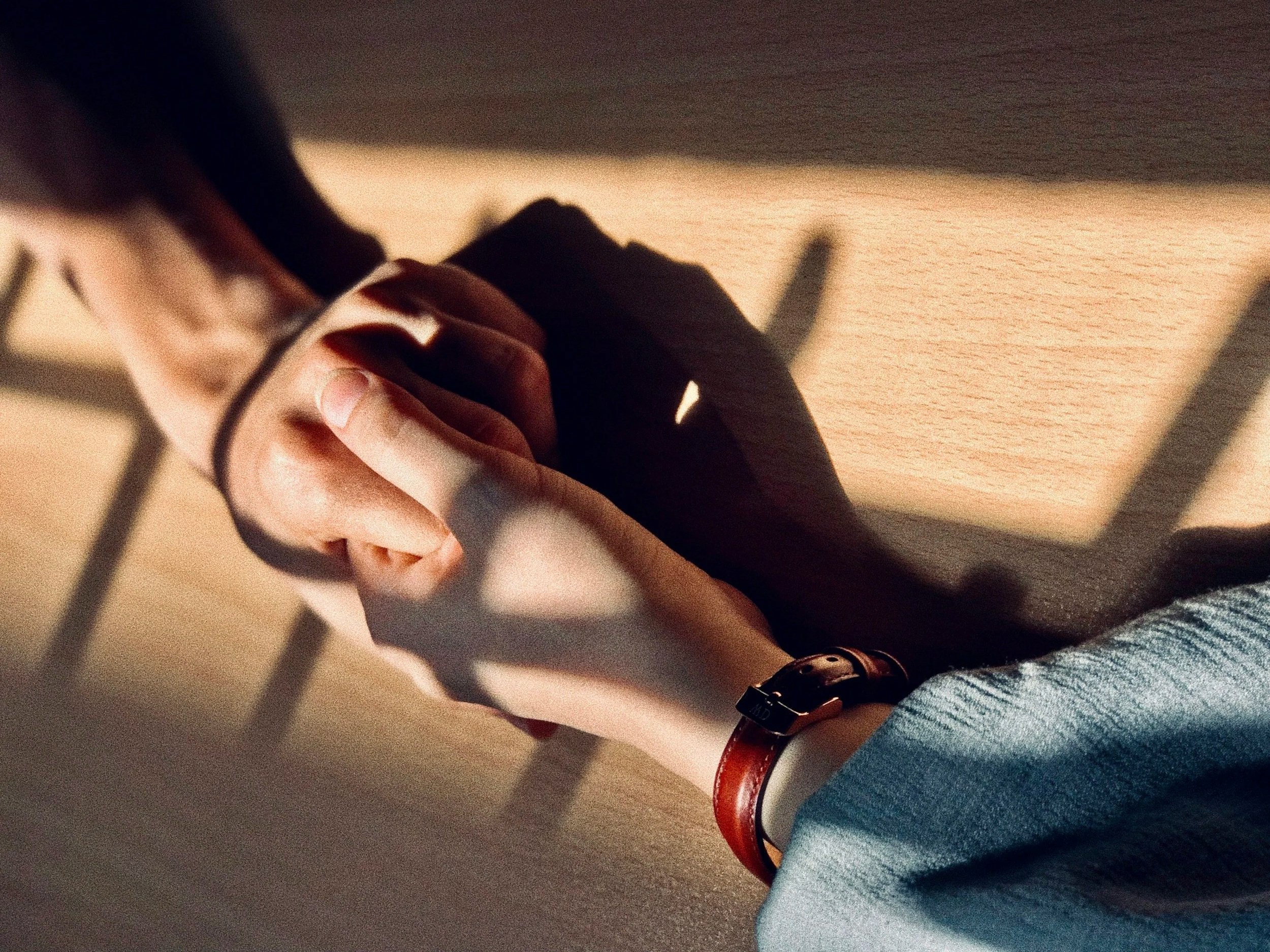 Close-up of two people holding hands on a wooden table with shadows from window blinds.