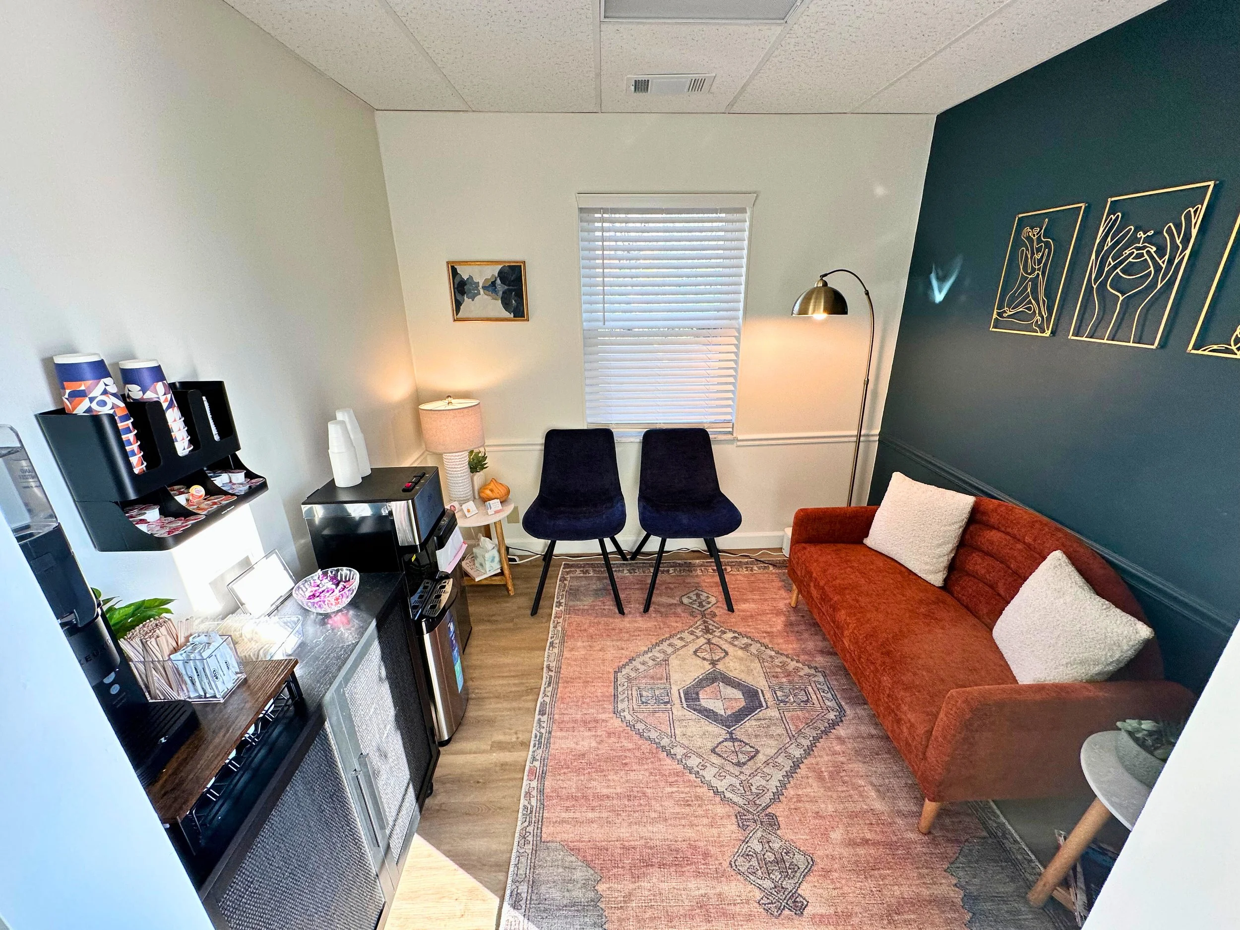 A cozy sitting room with a brick orange sofa, two black chairs, a vintage-style area rug, a side table with a lamp, and a small table with a plant. There is a coffee station with a microwave, coffee dispenser, cups, and a water cooler. The wall art includes abstract line drawings. Sunshine enters through closed white blinds.