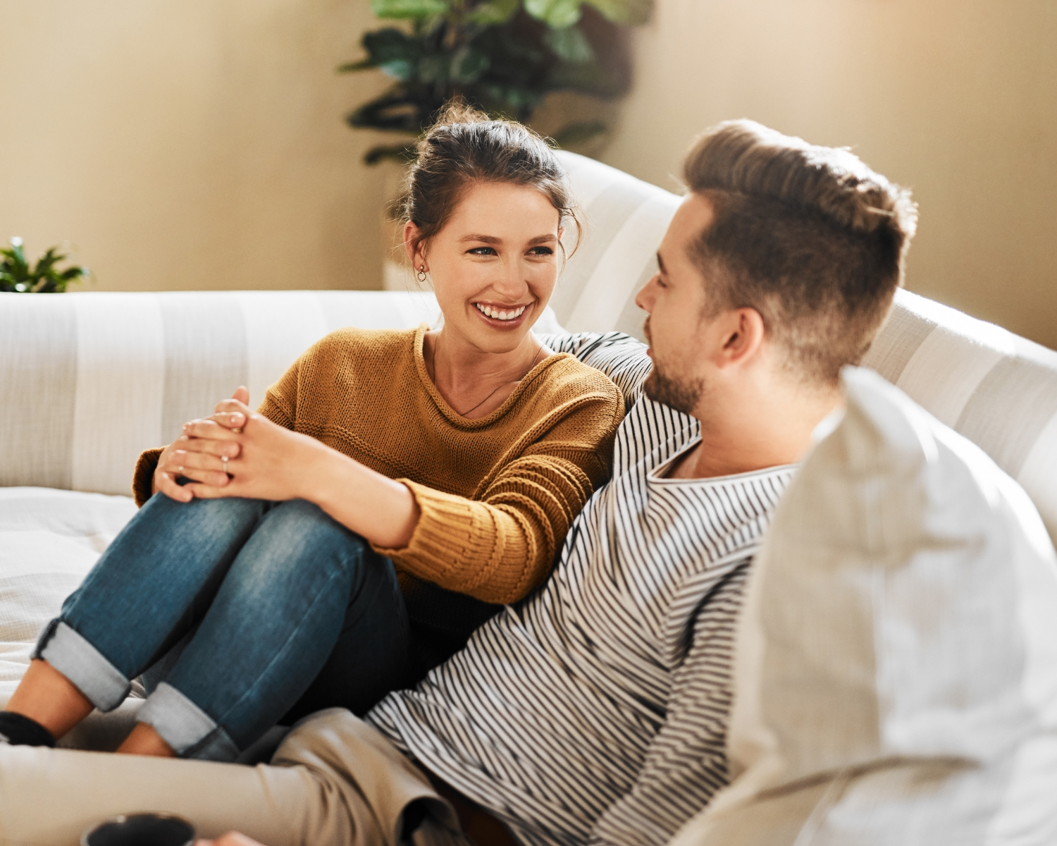 A woman and man sitting on a cream-colored couch, smiling and engaging in conversation indoors with sunlight, plants, and neutral decor in the background.