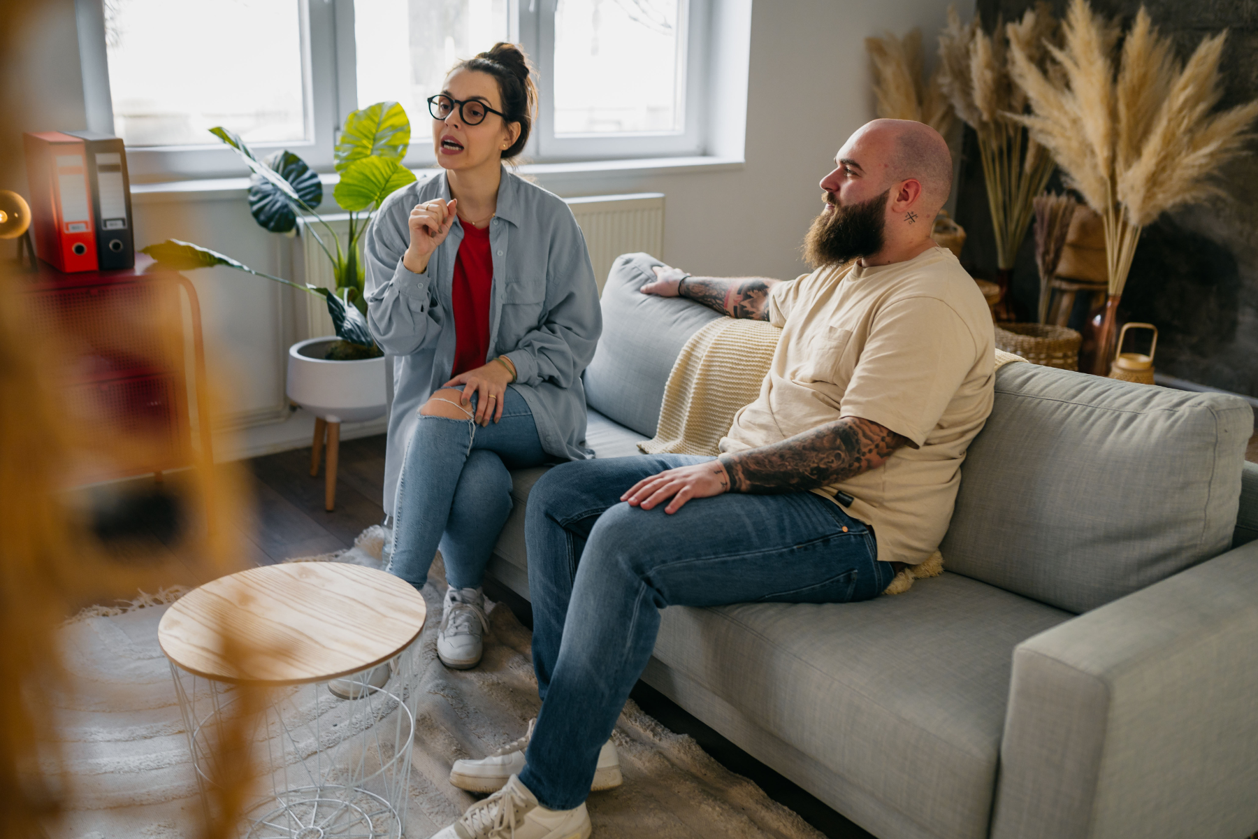 A woman with glasses and a man with a beard having a conversation in a living room with houseplants, a sofa, and decorative dried plants.