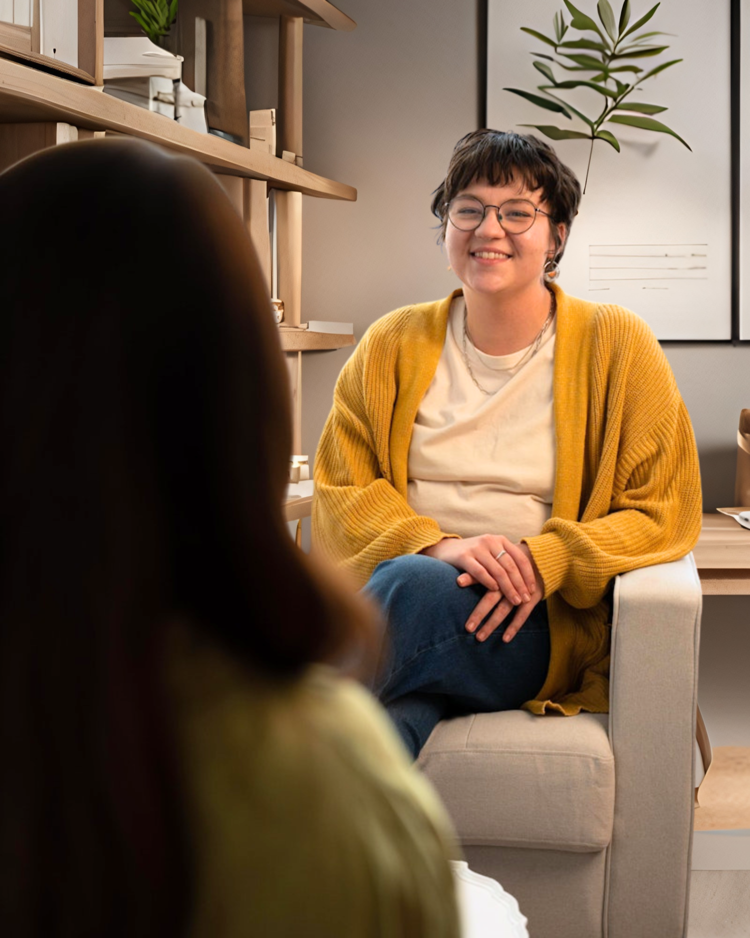 A woman with short dark hair, glasses, and earrings sitting comfortably on a beige armchair, smiling and wearing a yellow cardigan over a white t-shirt, in an office or therapy room with shelving and artwork in the background.