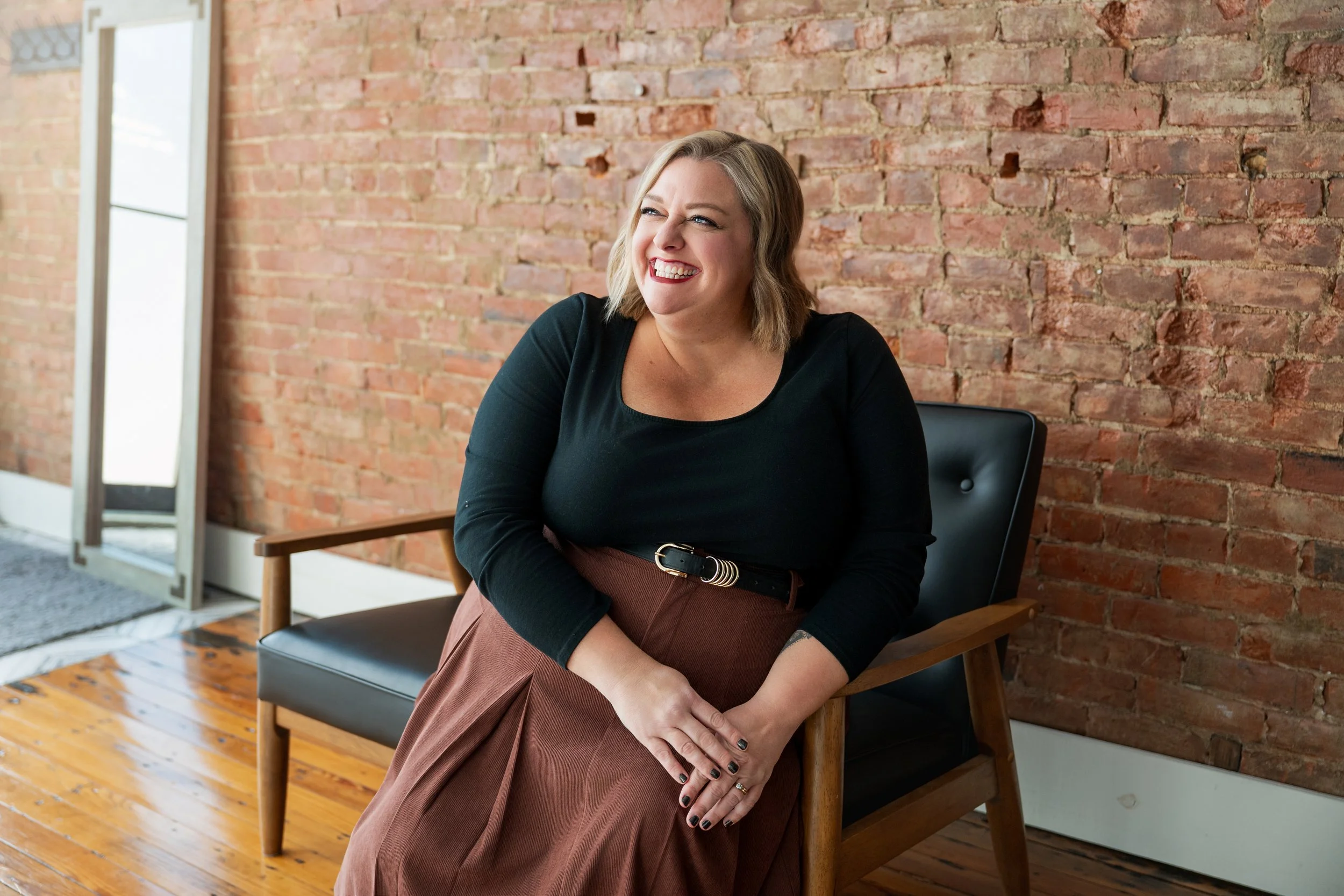 Woman sitting on a black and wooden chair, smiling and looking to the side inside a room with a brick wall background.