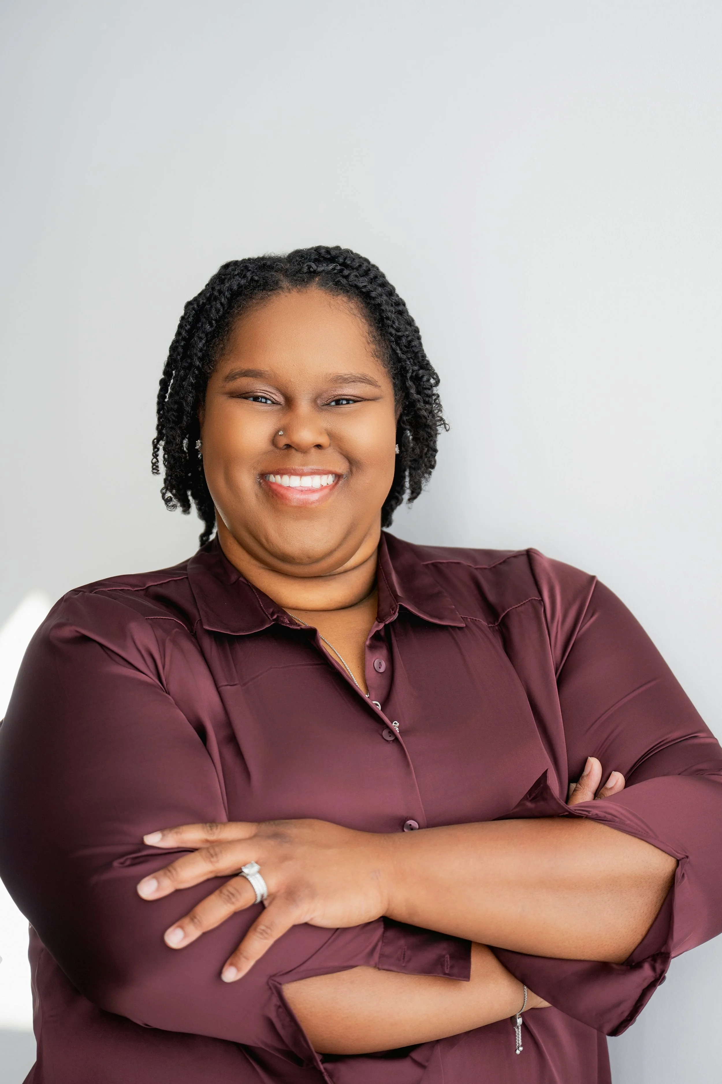 Portrait of a smiling woman with crossed arms, wearing a burgundy blouse, standing against a plain background.