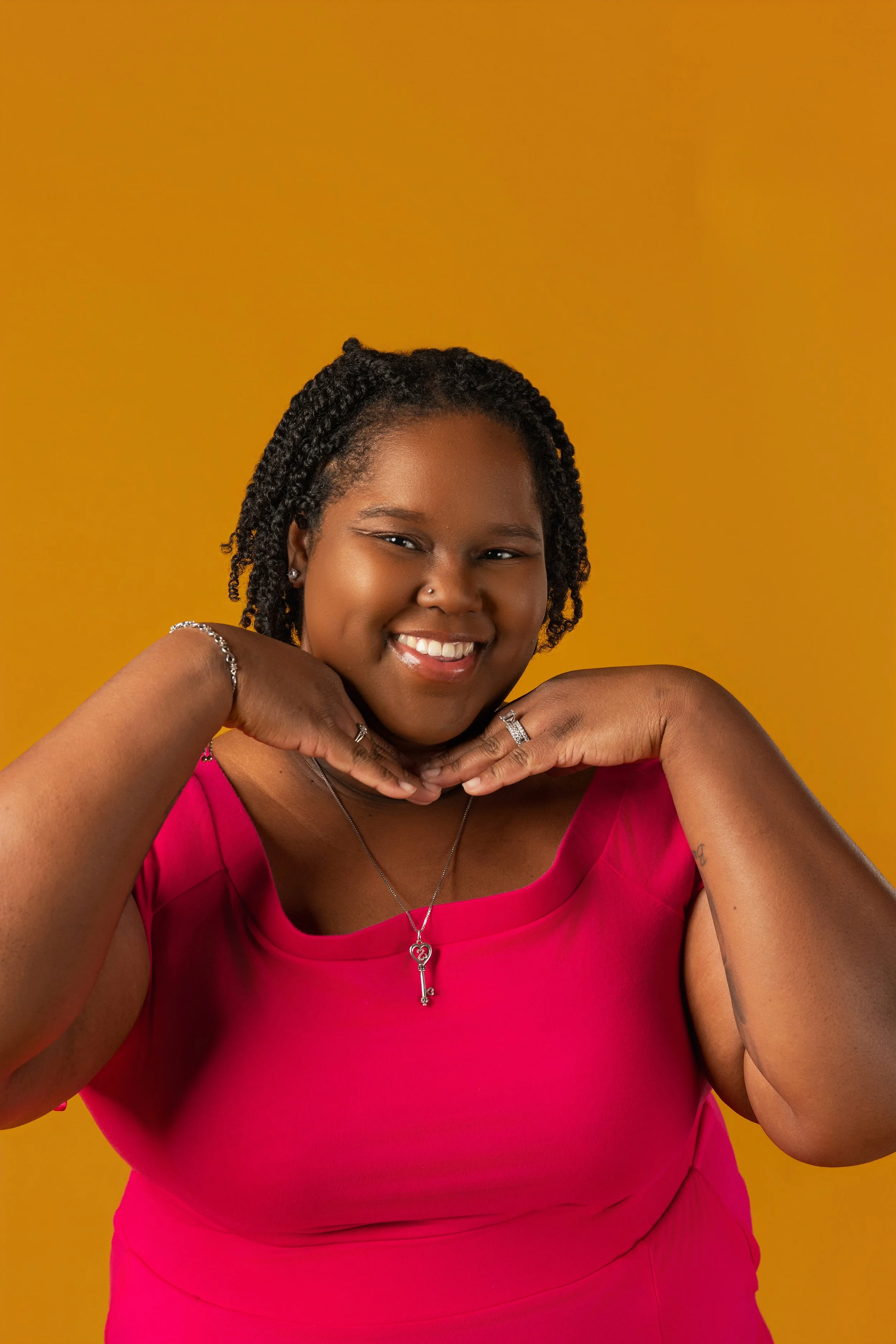 A young woman with dark curly hair, smiling and wearing a bright pink top, posed with her hands under her chin against a yellow background.
