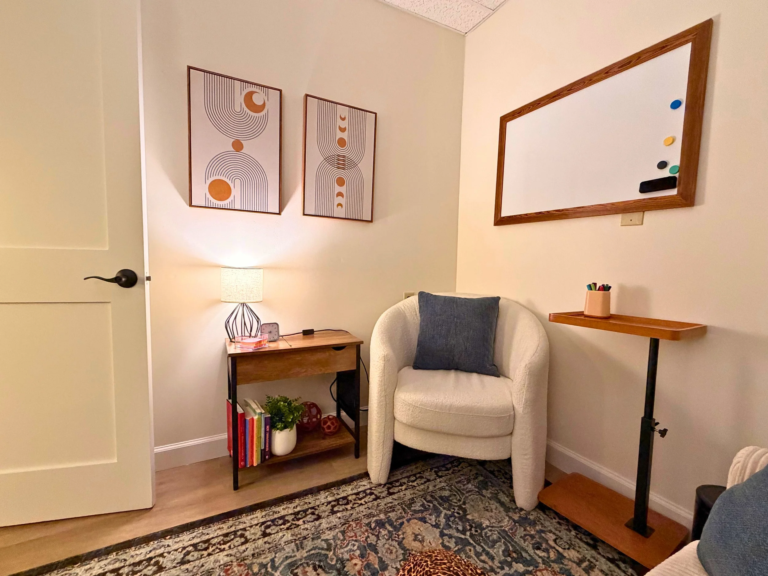 Cozy reading nook with a white armchair that has a dark gray cushion, a small side table with books and a plant, a modern desk lamp, abstract wall art, and a wooden-framed whiteboard with magnets, in a room with a patterned rug.
