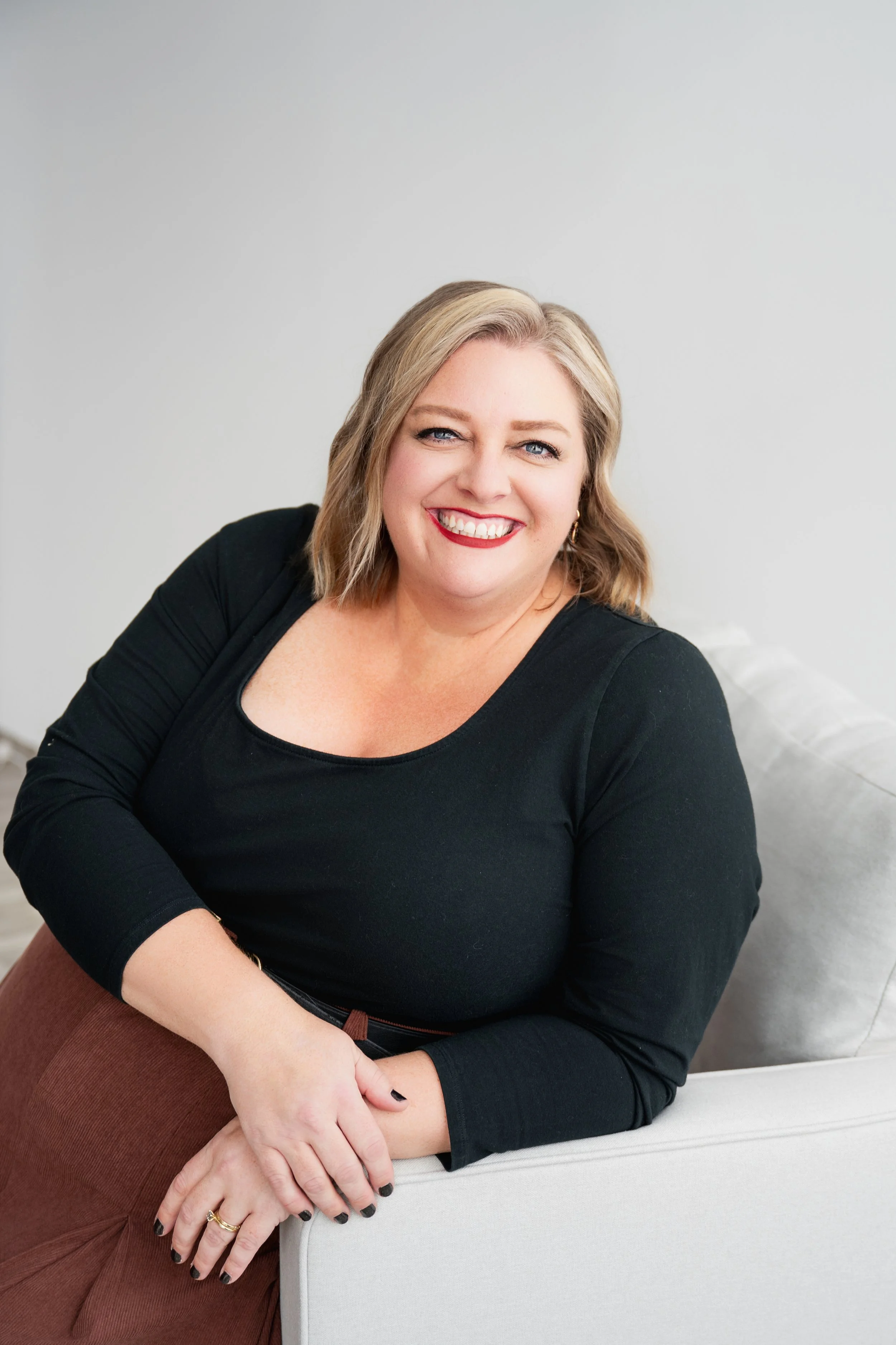 A woman with blonde hair, wearing a black top, sitting on a grey sofa, smiling at the camera.