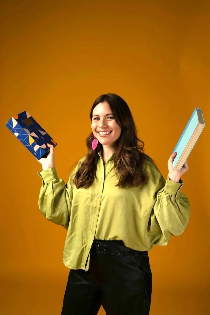 A woman with long brown hair, wearing a yellow blouse and black pants, smiling and holding a colorful patterned notebook in her right hand and a light blue book in her left hand, standing against a solid orange background.