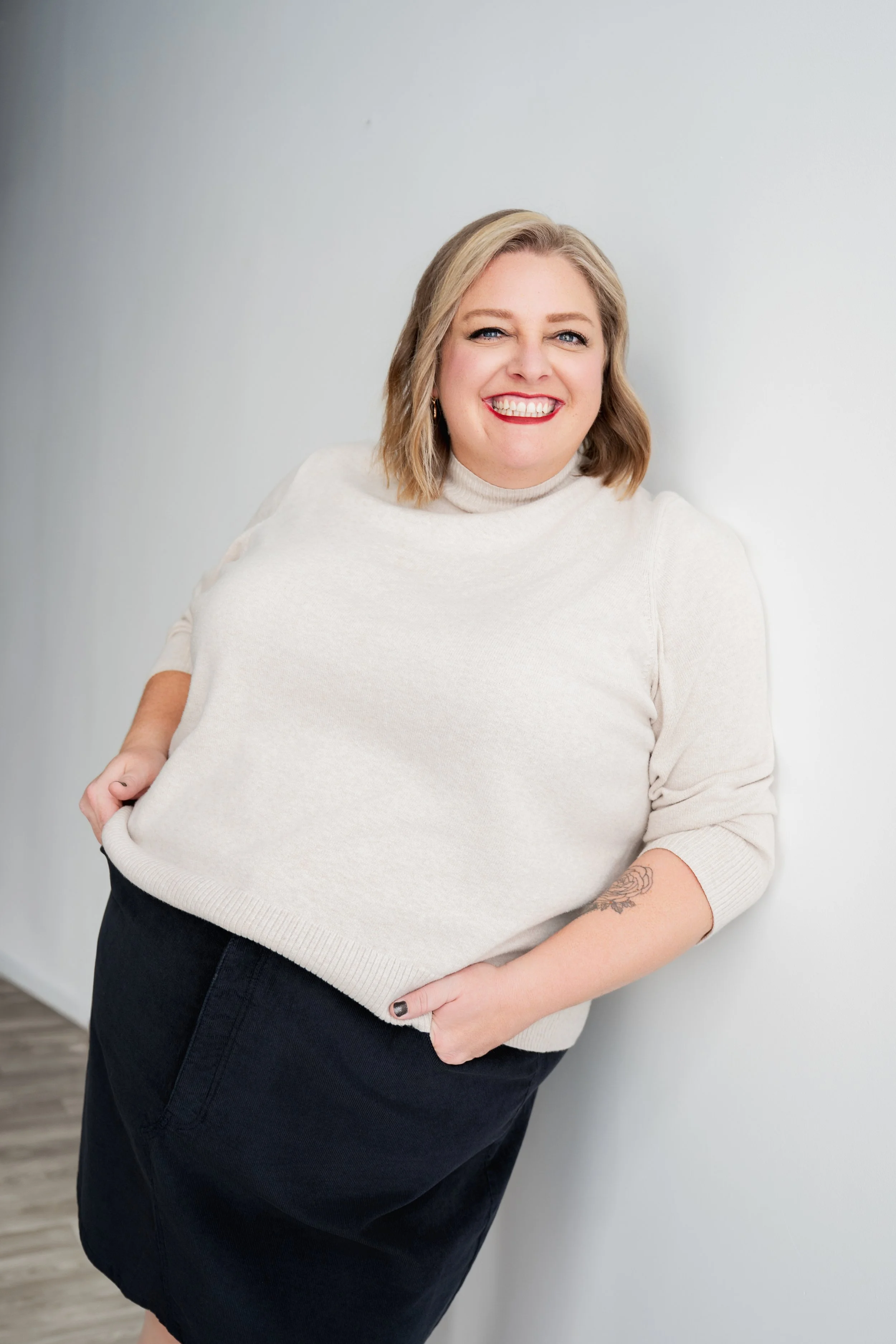 A smiling woman with blonde hair, wearing a light-colored sweater and dark skirt, standing against a plain, light-colored wall.