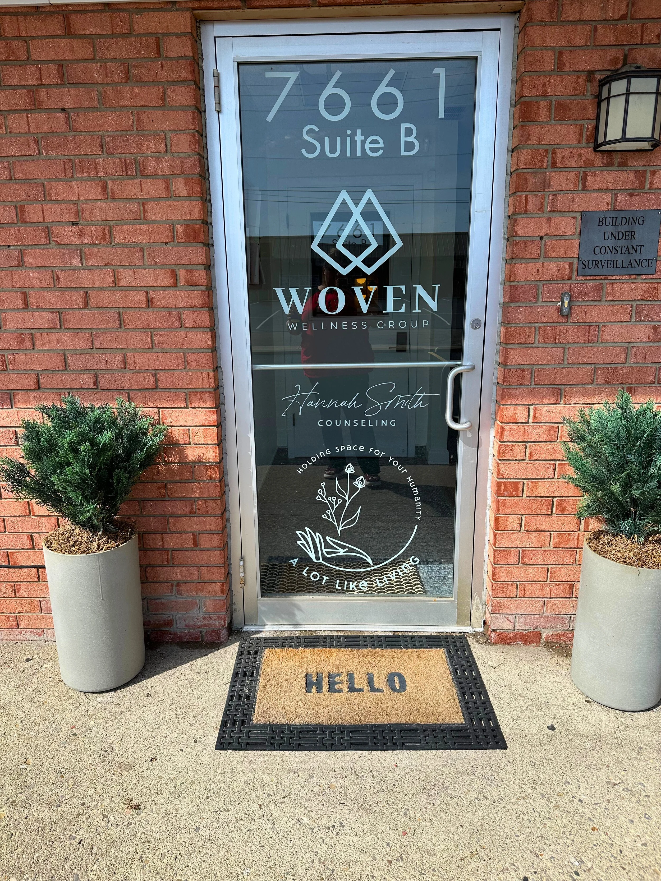 Front door of a wellness counseling business with the address 7661 Suite B, the business name Woven Wellness Group, and Hannah Smith Counseling written on the glass door. There are two potted plants on either side of the door and a black and tan doormat with 'HELLO' written on it.