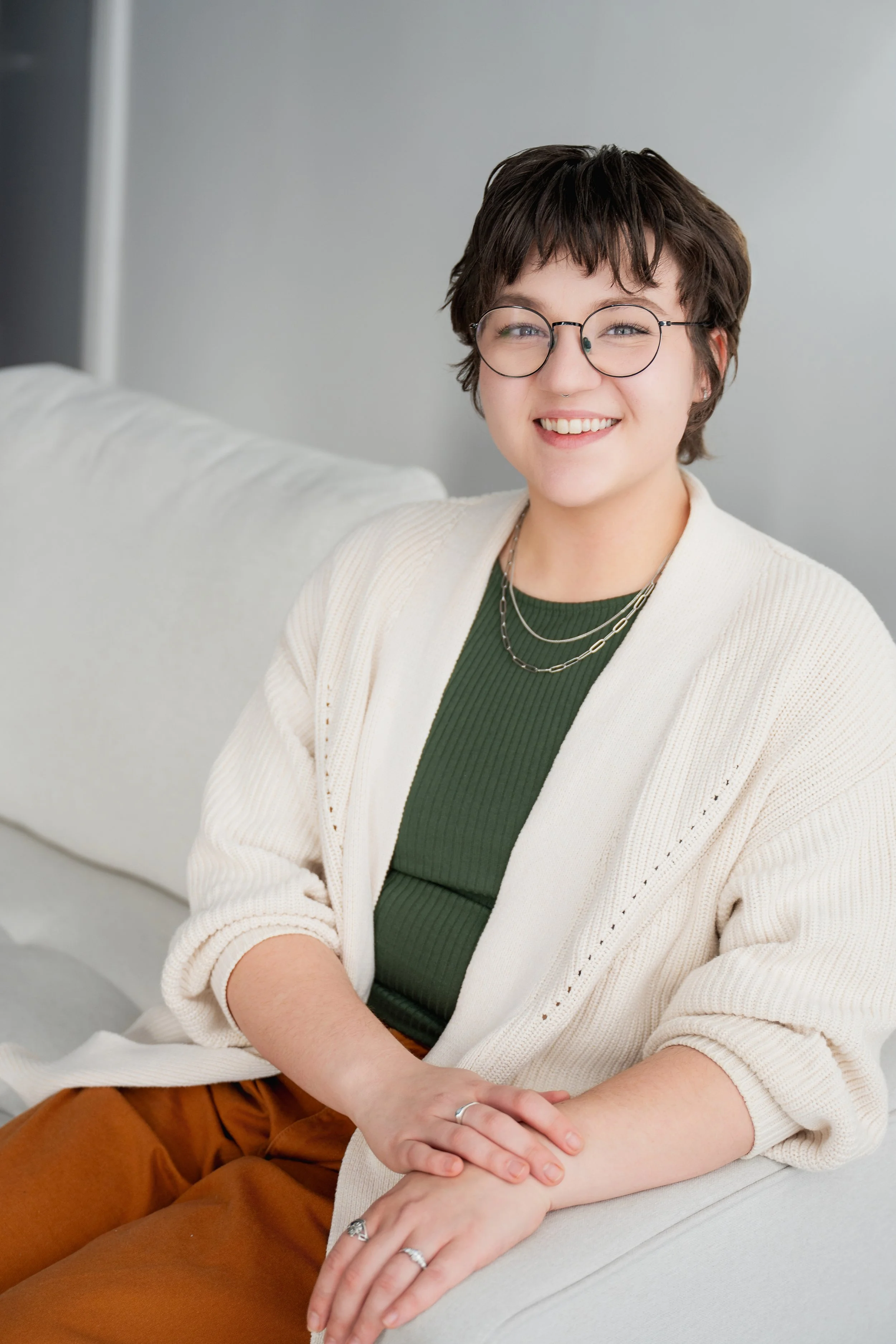 A young woman with short dark hair, glasses, and a big smile sitting on a light-colored sofa. She is wearing a green ribbed top, layered necklaces, a white cardigan, and brown pants.