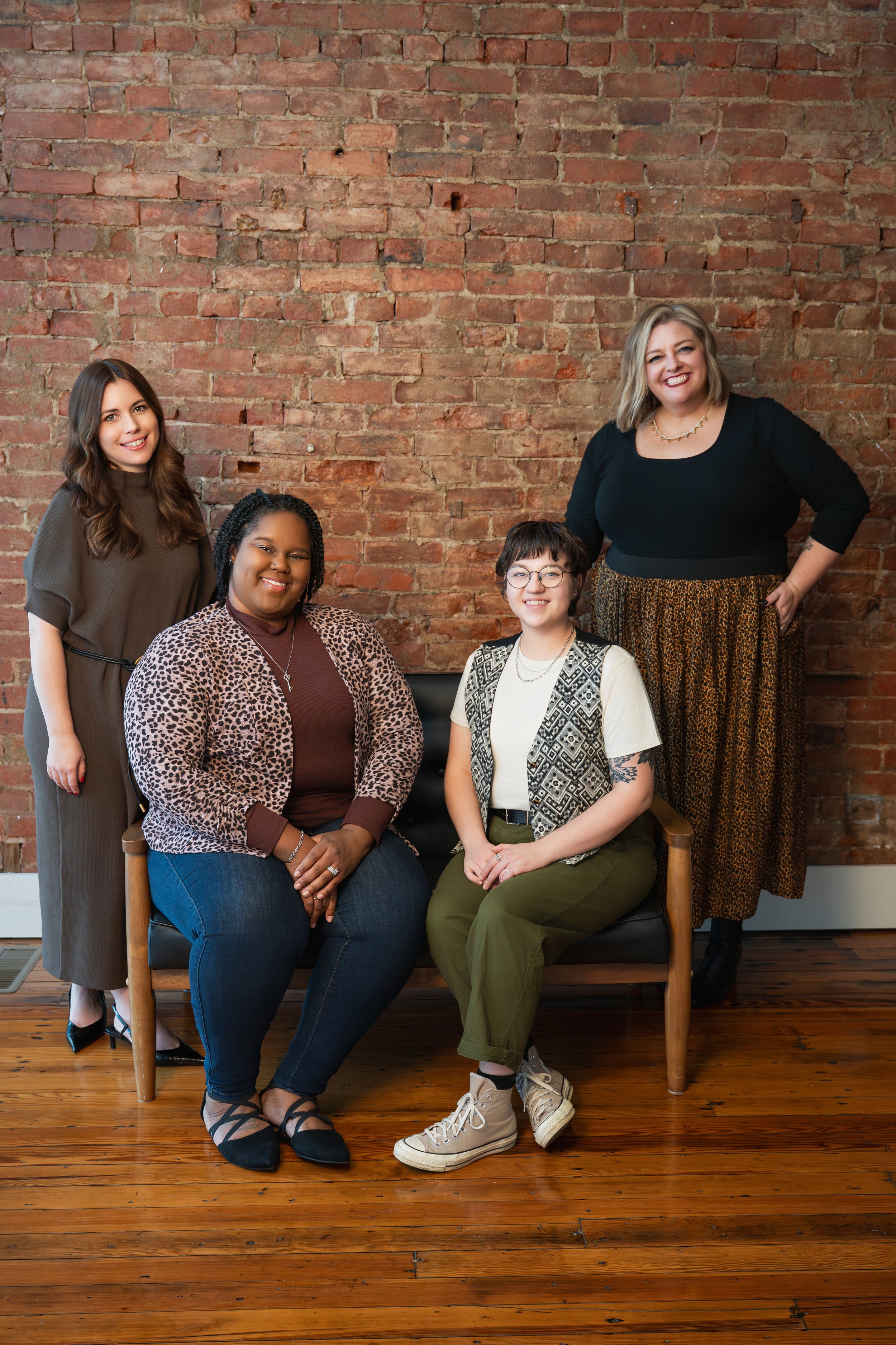 Four women posing together indoors against a brick wall, with two seated on a black sofa and two standing behind them.