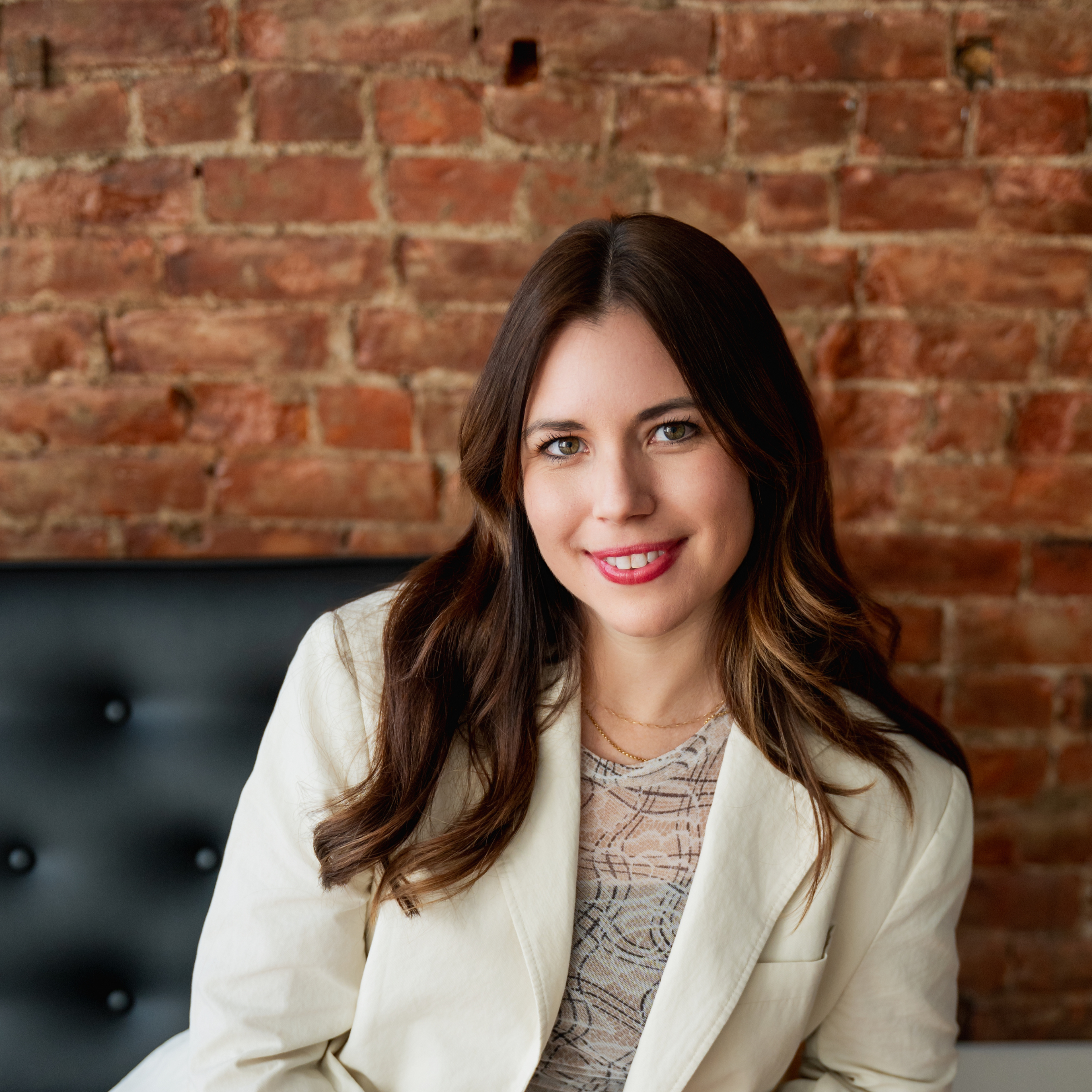 A woman with long brown hair and light-colored eyes smiling at camera, wearing a cream blazer over a patterned top, sitting in front of a brick wall.
