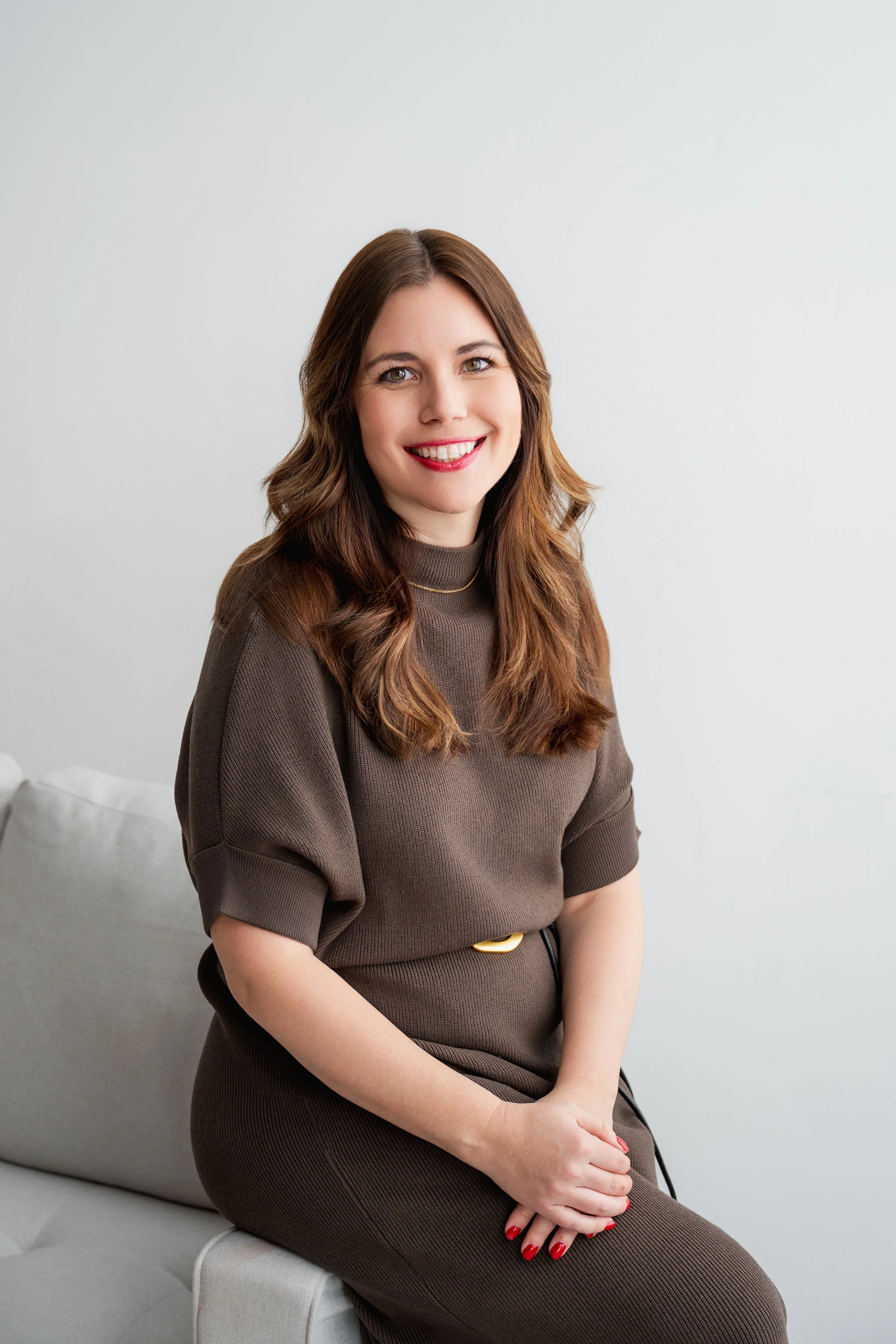 A woman with long, wavy brown hair, smiling, wearing a brown dress, sitting on a light-colored sofa against a plain white wall.