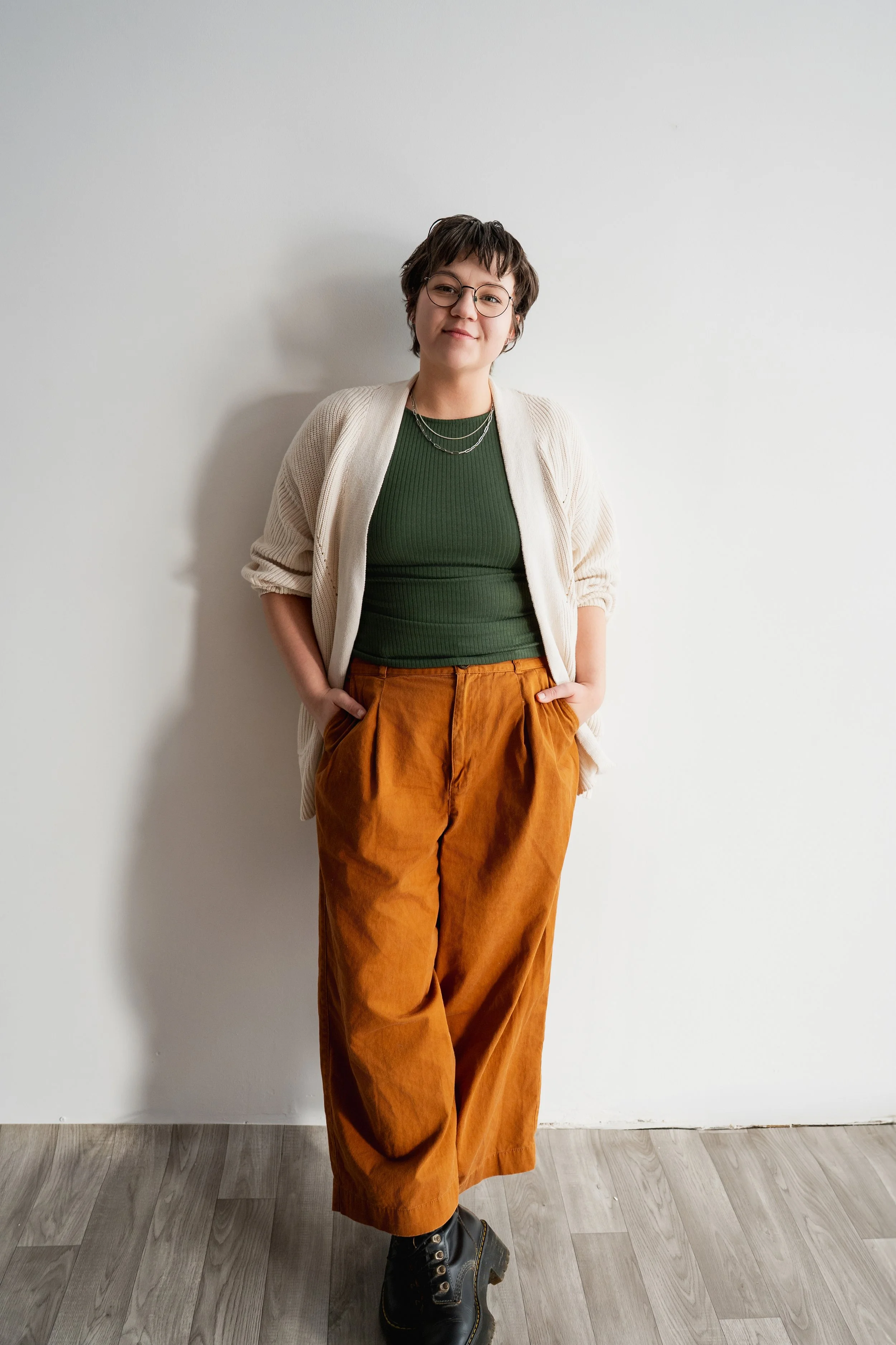A woman with short dark hair and glasses standing against a plain white wall, wearing a green top, beige cardigan, orange pants, and black boots, with her hands in her pockets.