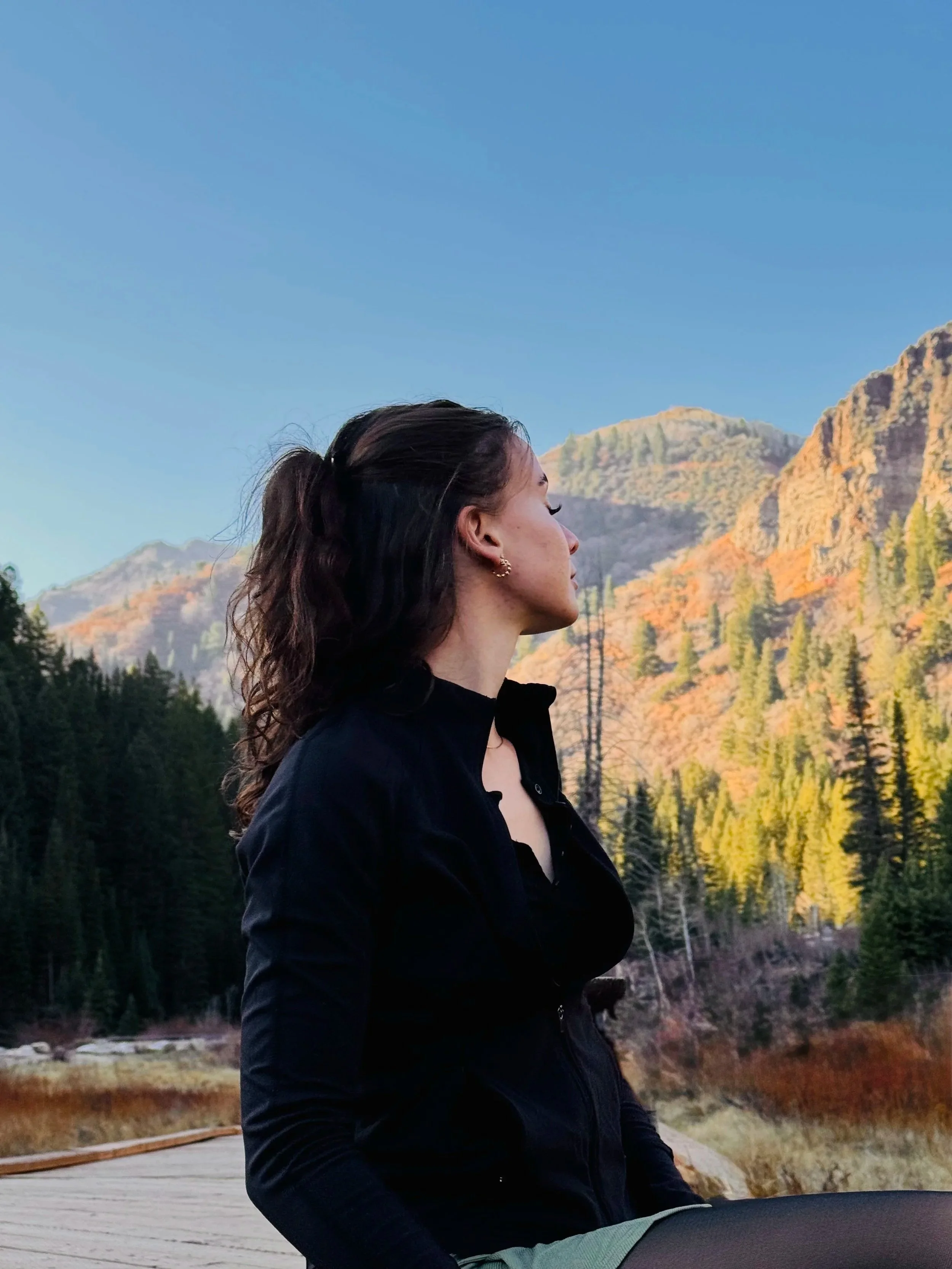 A woman with dark, wavy hair tied back in a ponytail, wearing a black jacket, is sitting outdoors with her eyes closed, facing a mountain landscape with autumn-colored trees, under a clear blue sky.