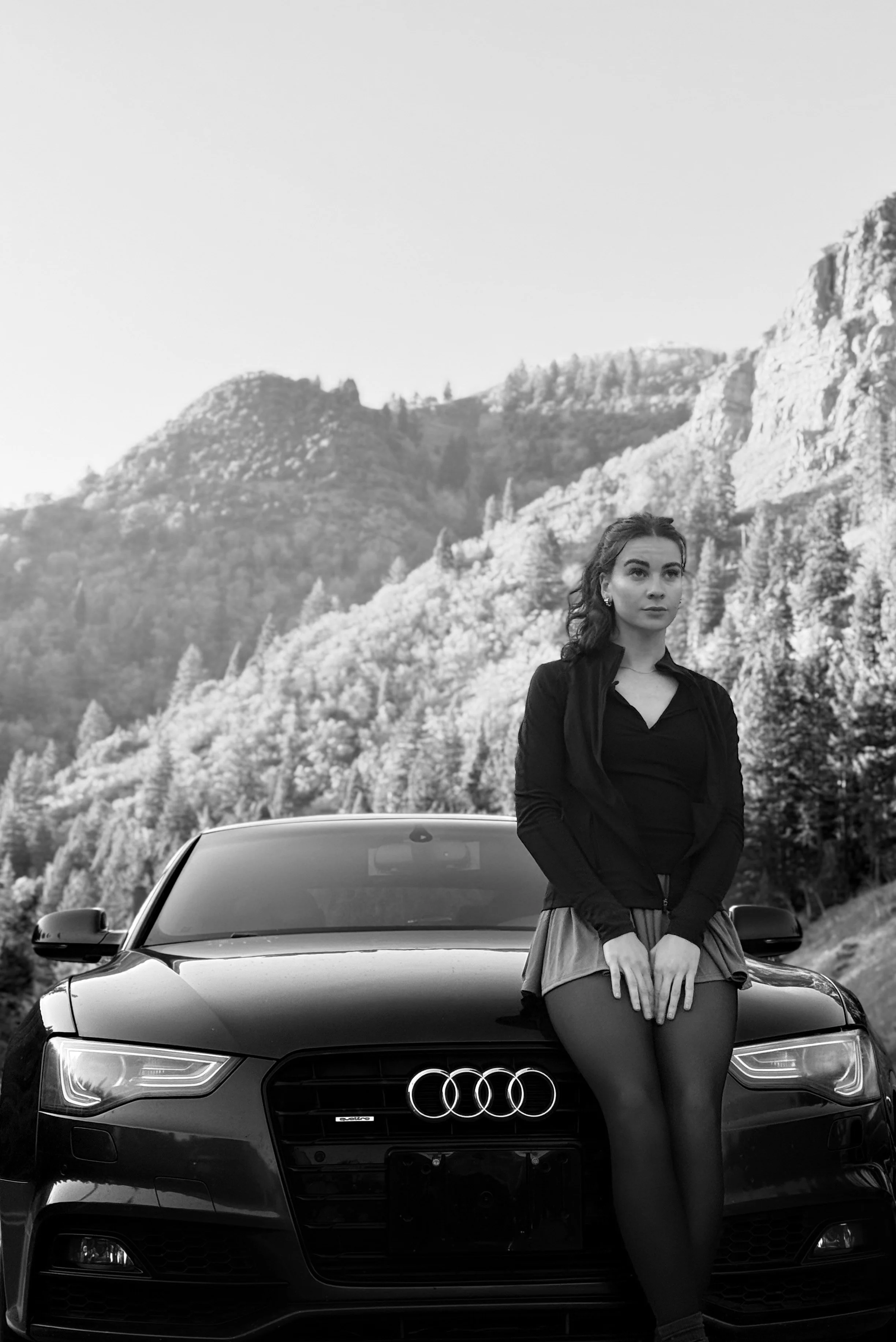 A woman standing in front of a black Audi car with mountains and trees in the background, black and white photo.