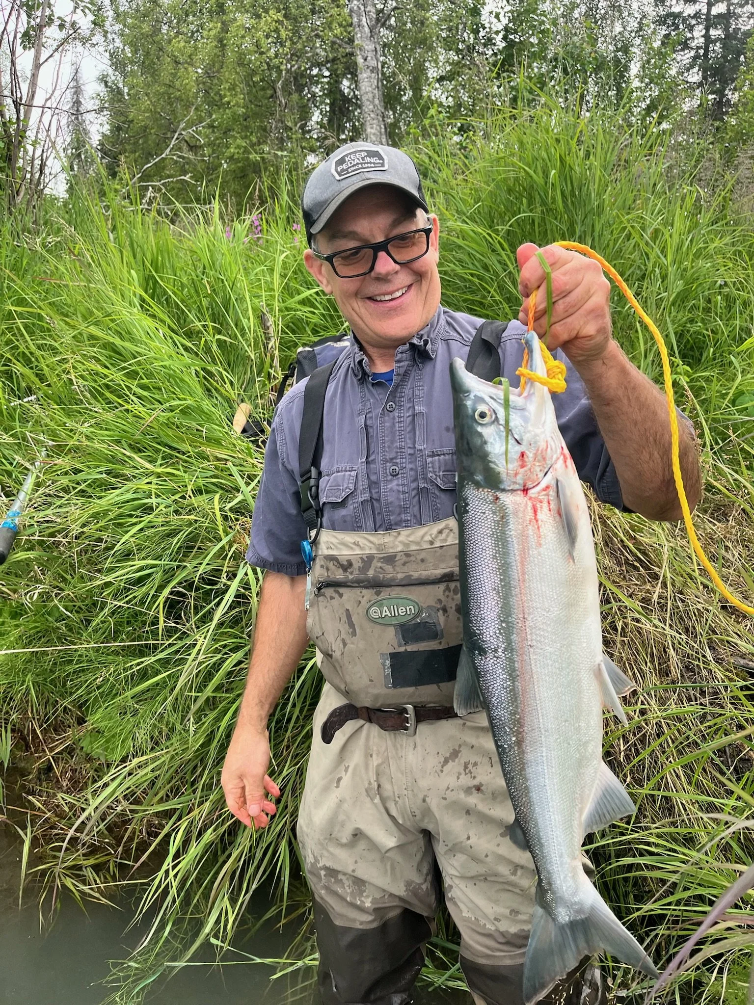 Smiling man in outdoor gear holding a large fish with a yellow string, standing in a green wetland area with tall grass and trees.