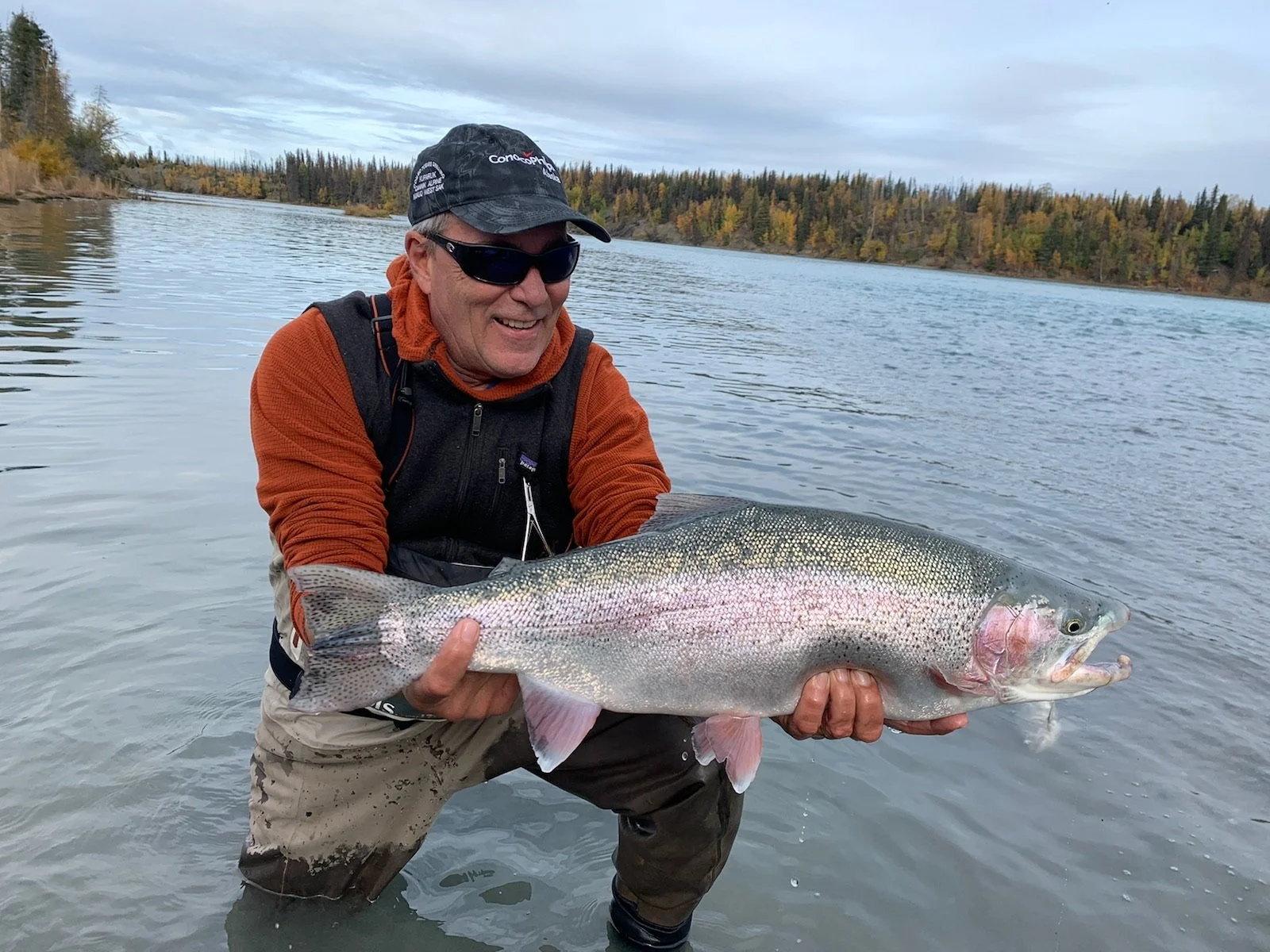 Man wearing sunglasses, a black cap, orange sleeve jacket, and waders holding a large rainbow trout by a river with fall-colored trees in the background.