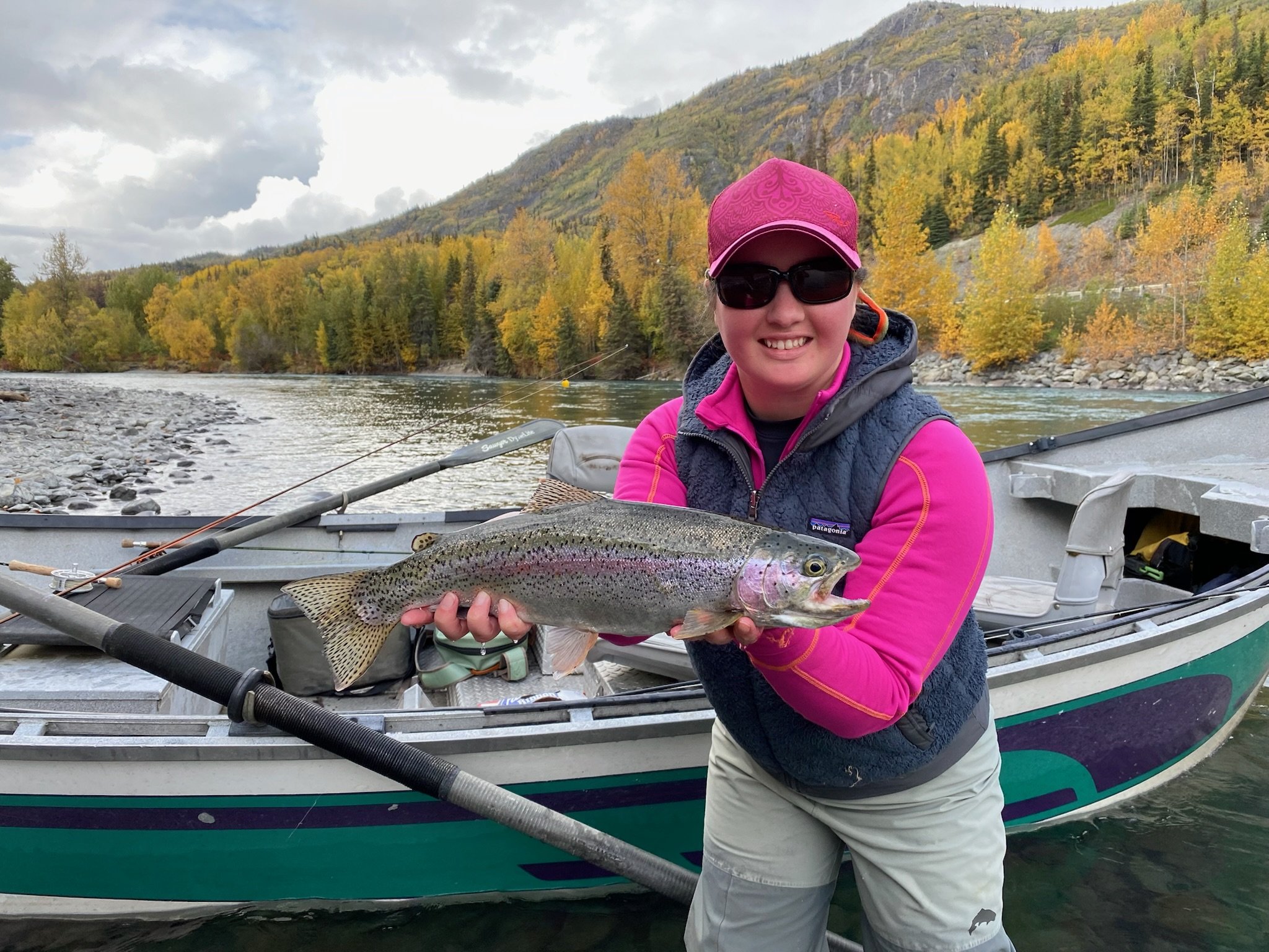 Woman wearing sunglasses, pink cap, and outdoor gear holding a large rainbow trout on a boat in a river with colorful fall trees and hills in the background.