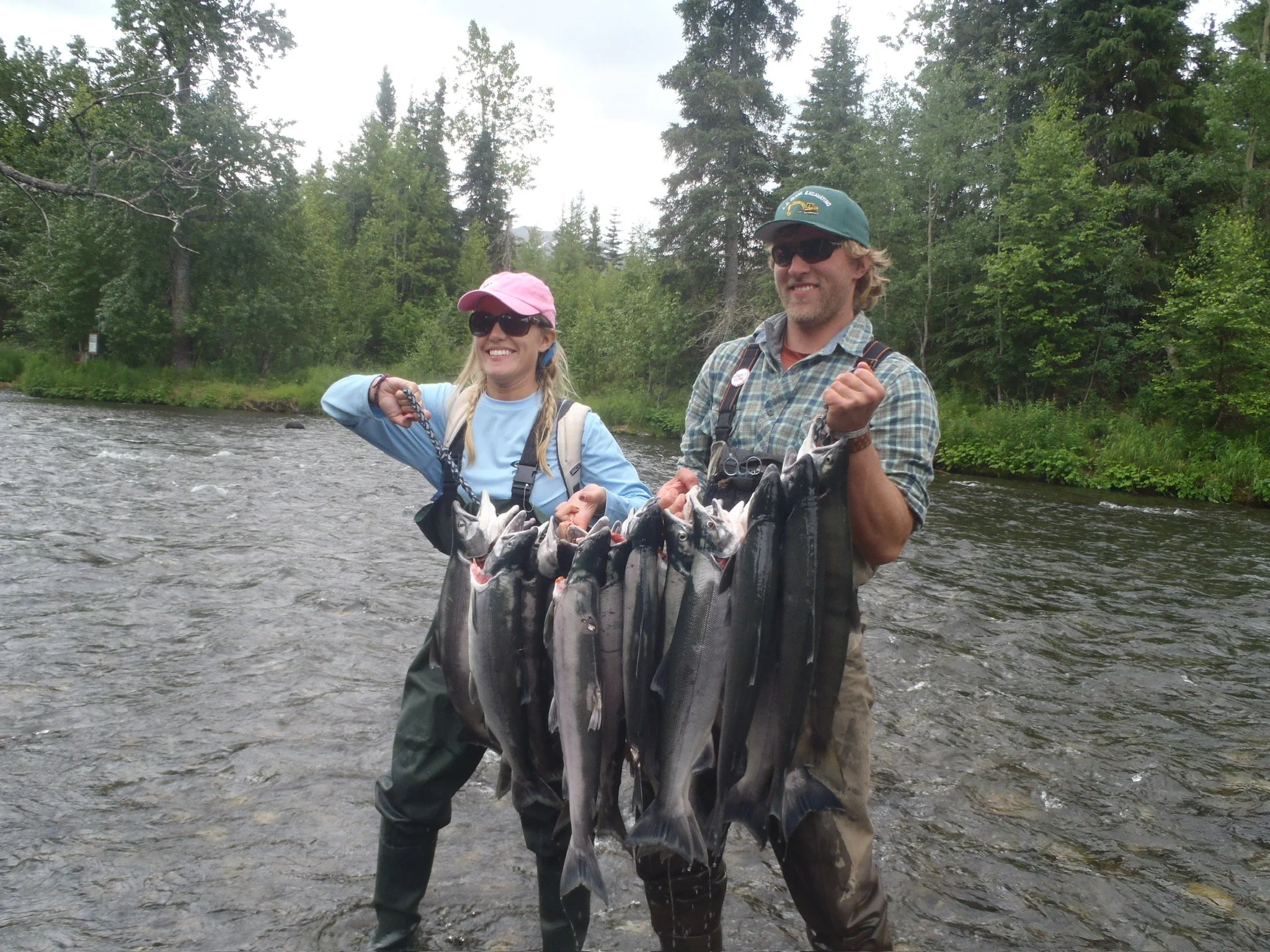 Two people in fishing gear holding a string of freshly caught fish in a river with a forest background.