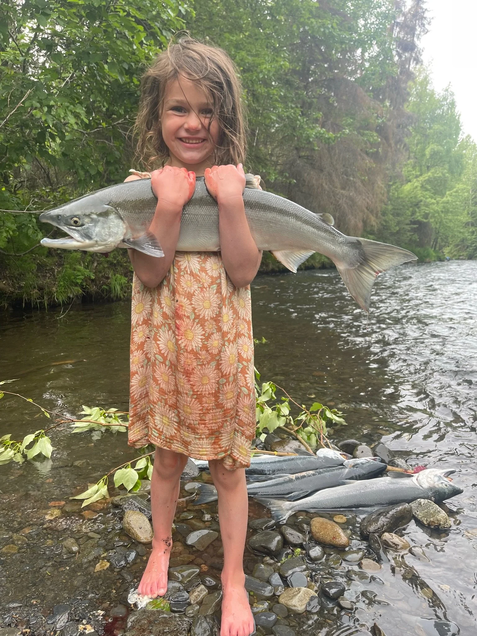Young girl standing barefoot near a river holding a large fish with a grin on her face, with several other fish on the rocks behind her.