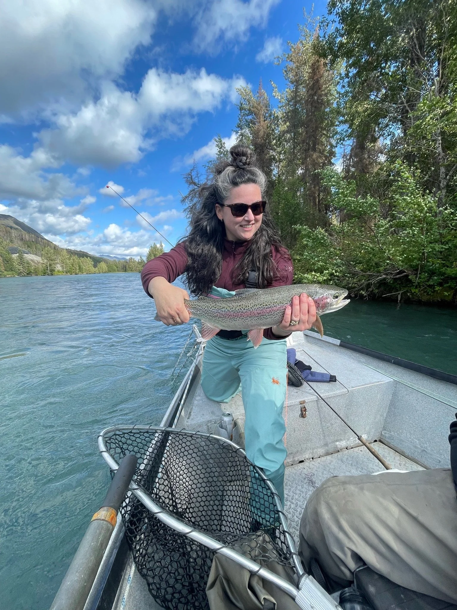 Woman with sunglasses and long curly hair holding a large rainbow trout on a boat in a river, with trees and blue sky in the background.