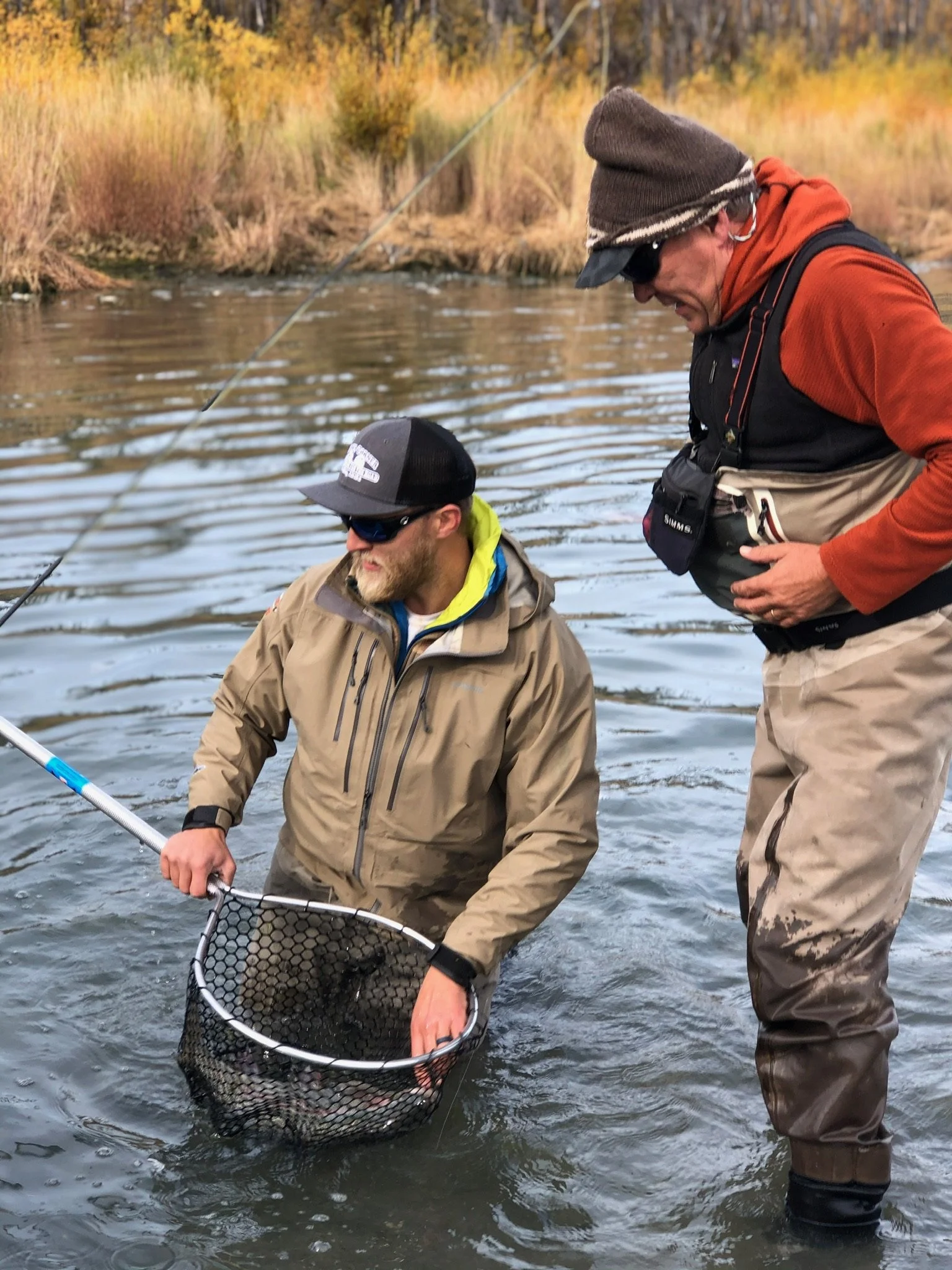Two men fishing in a river, one is holding a fishing net, and the other is standing close, wearing outdoor fishing gear and hats.