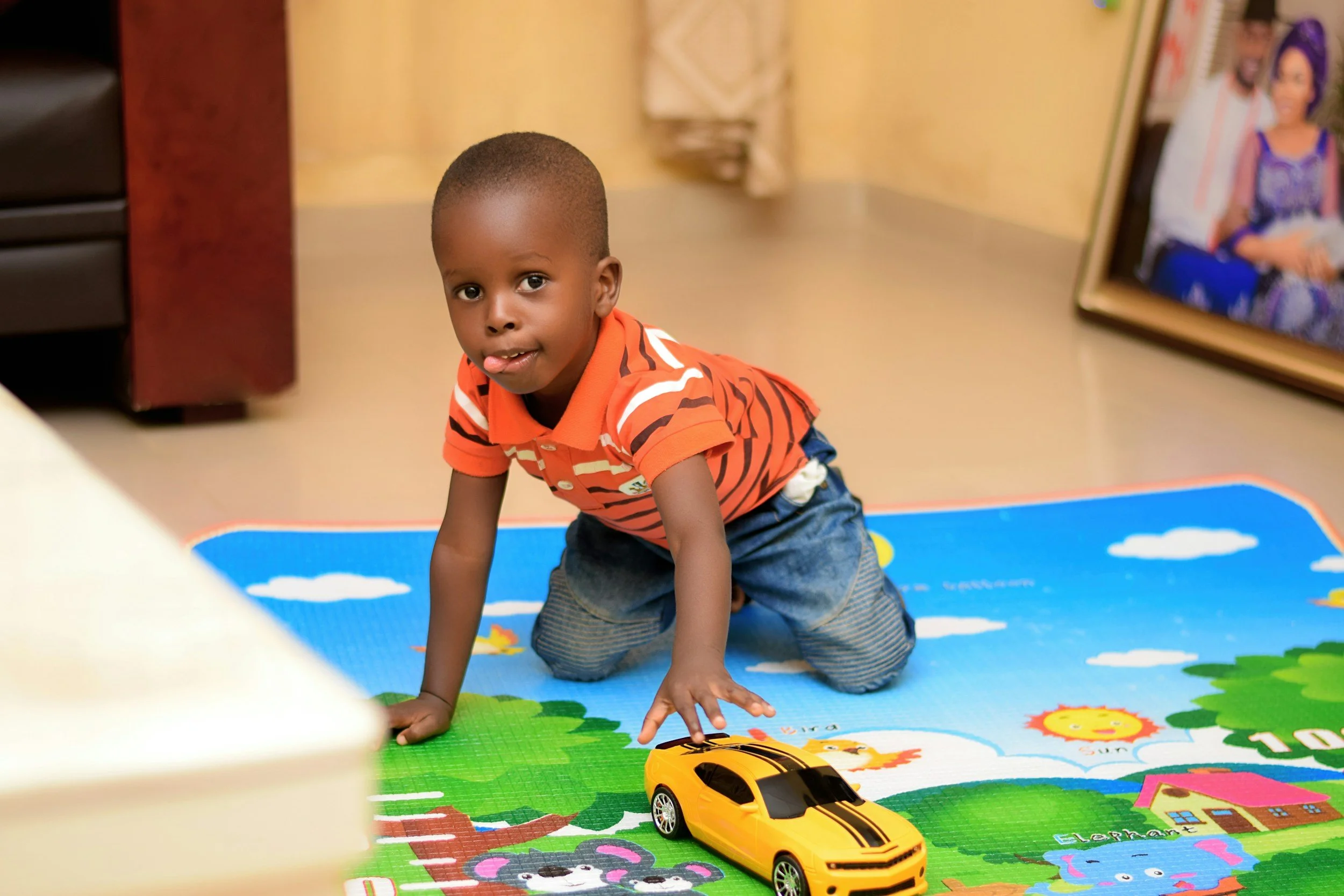 A young boy with a shaved head, wearing an orange striped polo shirt and jeans, is on his hands and knees on a colorful playground mat playing with a yellow toy car.
