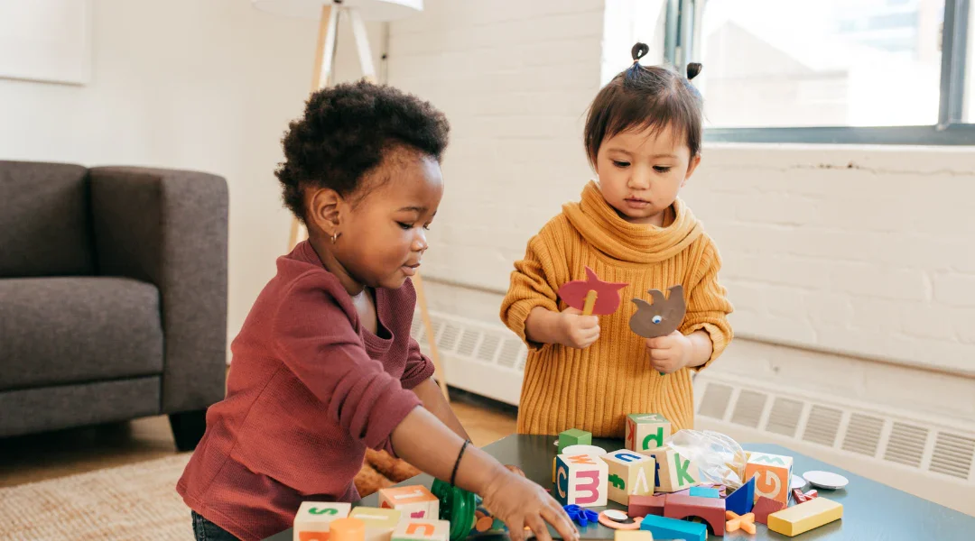 Two young girls playing with colorful wooden blocks and animal-shaped puppets on a table in a bright, cozy room.