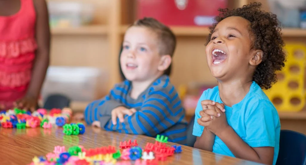 Two children sitting at a table with colorful alphabet puzzles, one child is laughing joyfully with hands clasped, the other is smiling in a classroom setting.