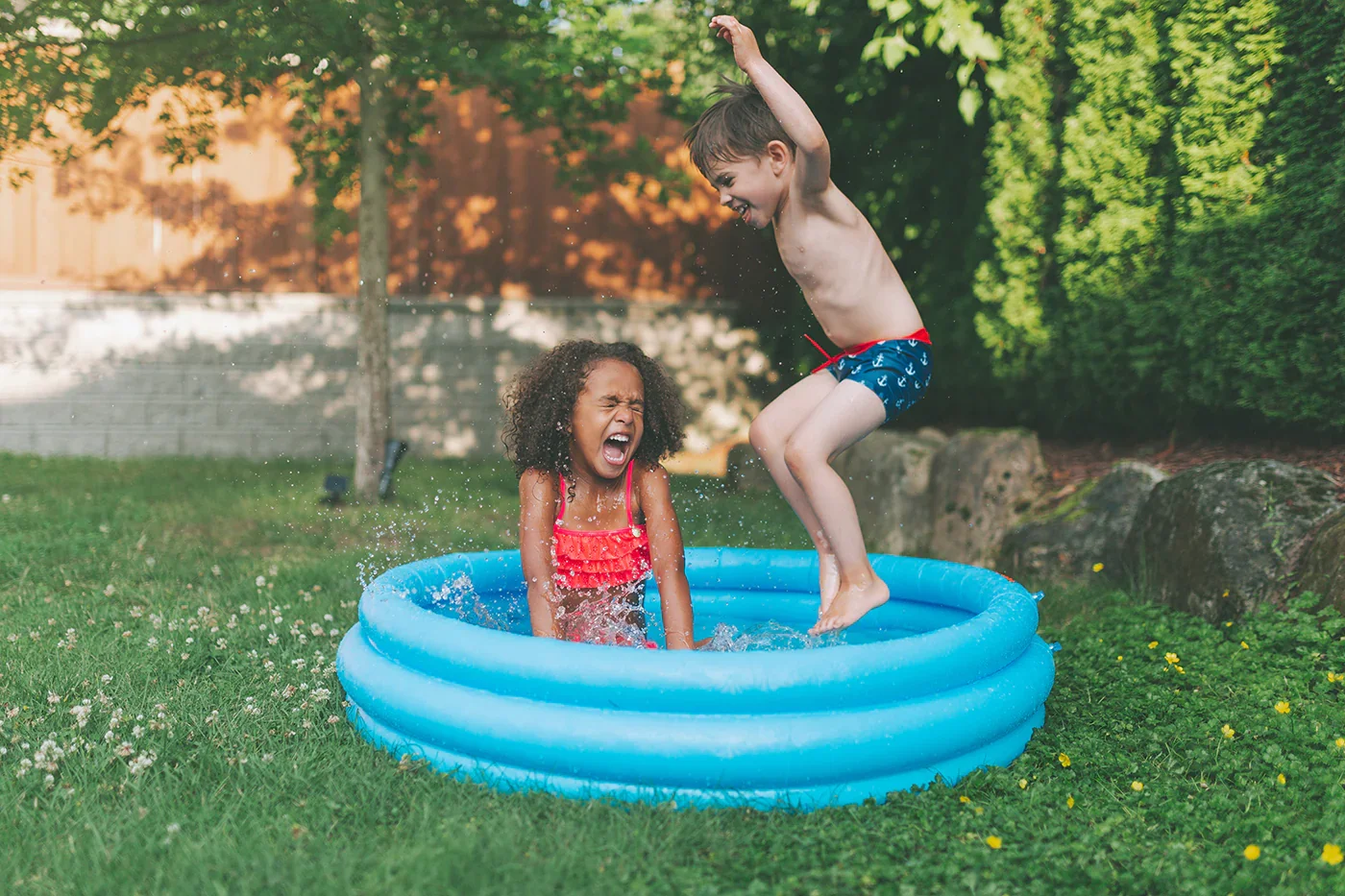Two children playing in a small inflatable pool outdoors, one girl sitting and laughing and one boy jumping into the pool, surrounded by grass and trees.