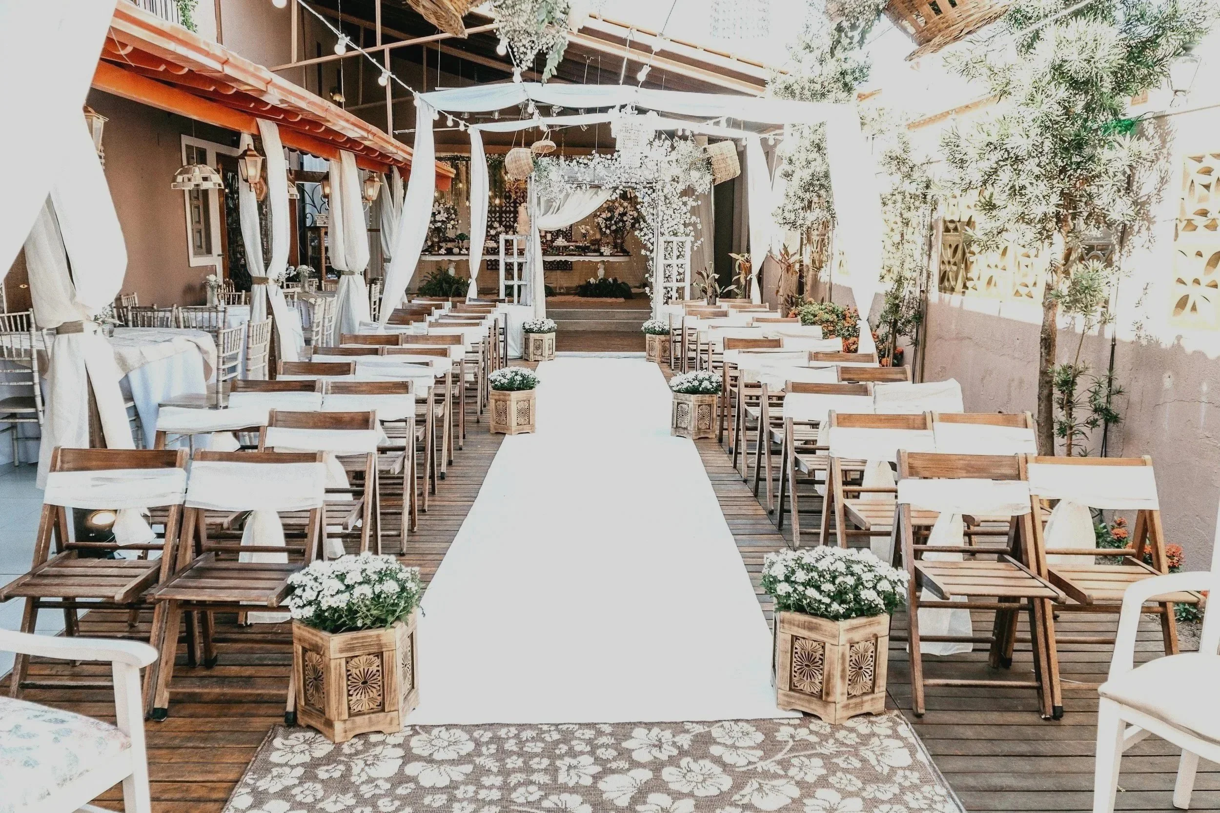 Wedding ceremony setup with rows of wooden chairs decorated with white fabric, a white aisle runner, floral arrangements in wooden planters, and a decorated arch at the front on a wooden deck patio.