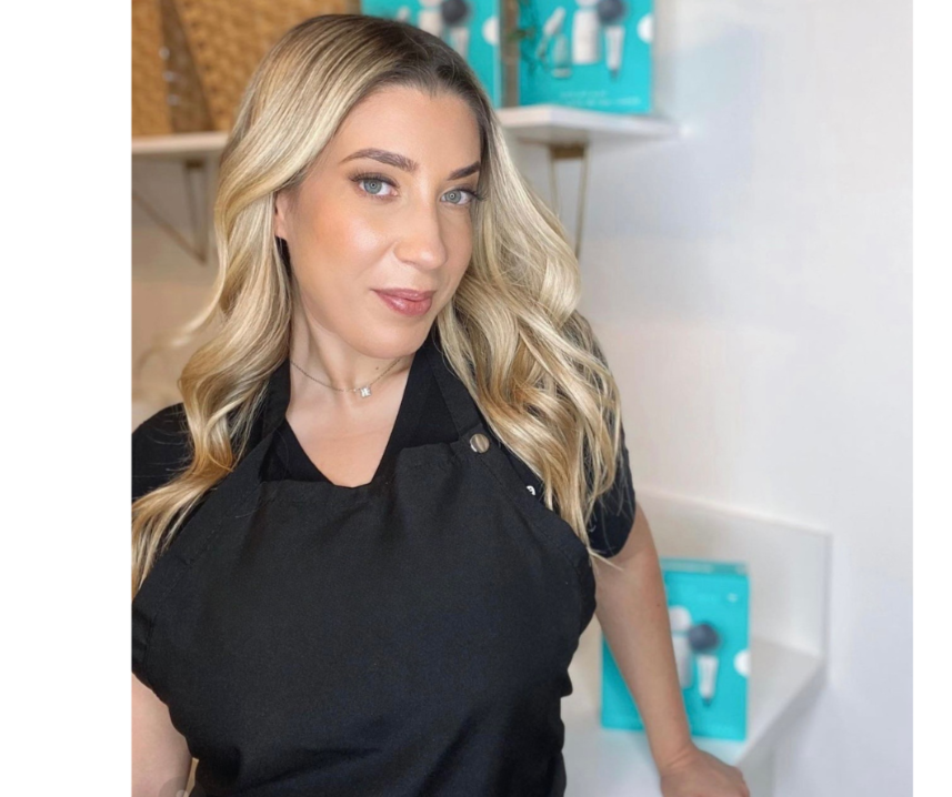 A woman with long, wavy blonde hair and blue eyes wearing a black apron in a bright kitchen or salon setting.