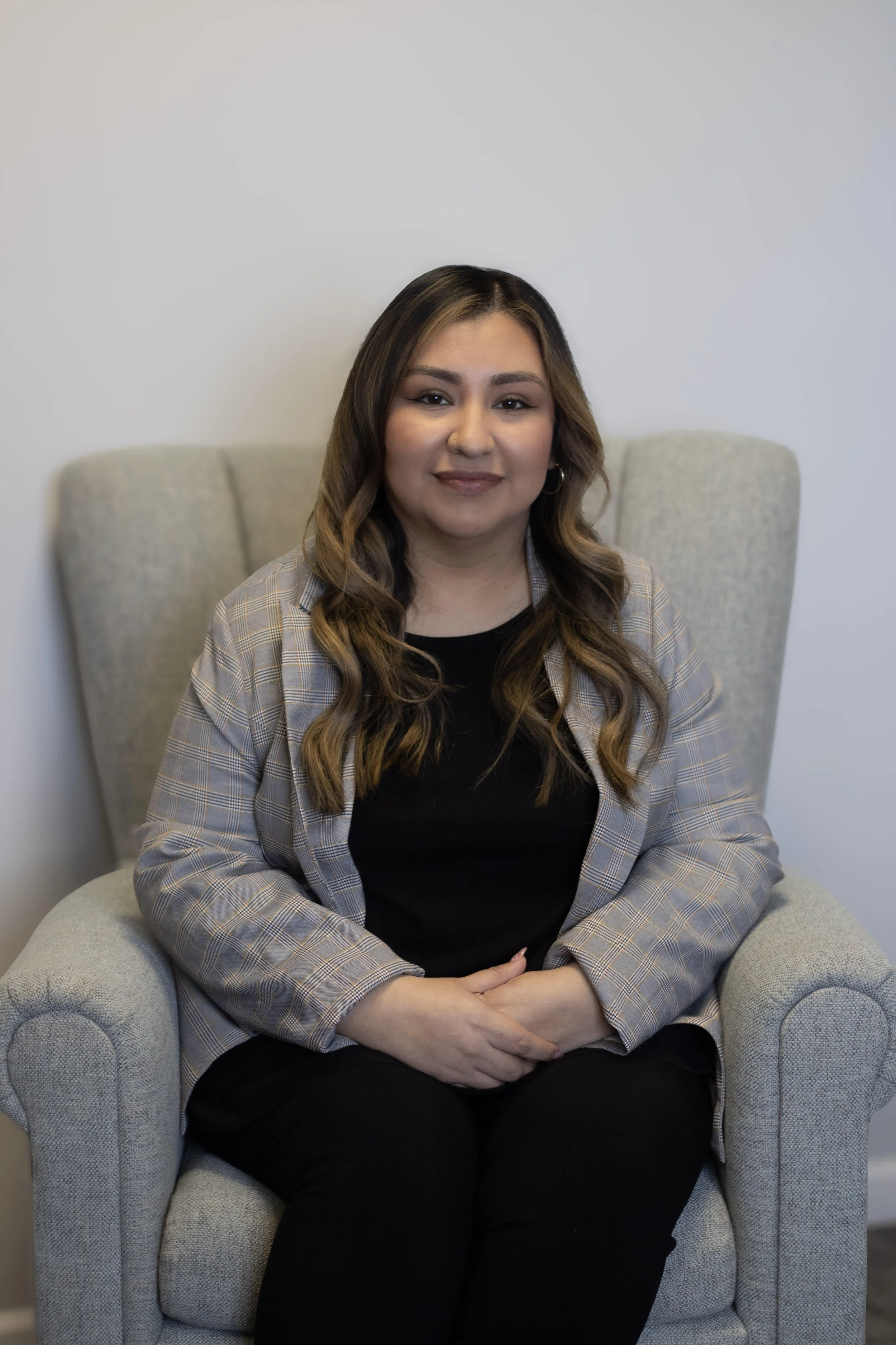 A woman with long wavy hair and hoop earrings, wearing a black top and plaid blazer, sitting in a light gray armchair against a plain white wall.