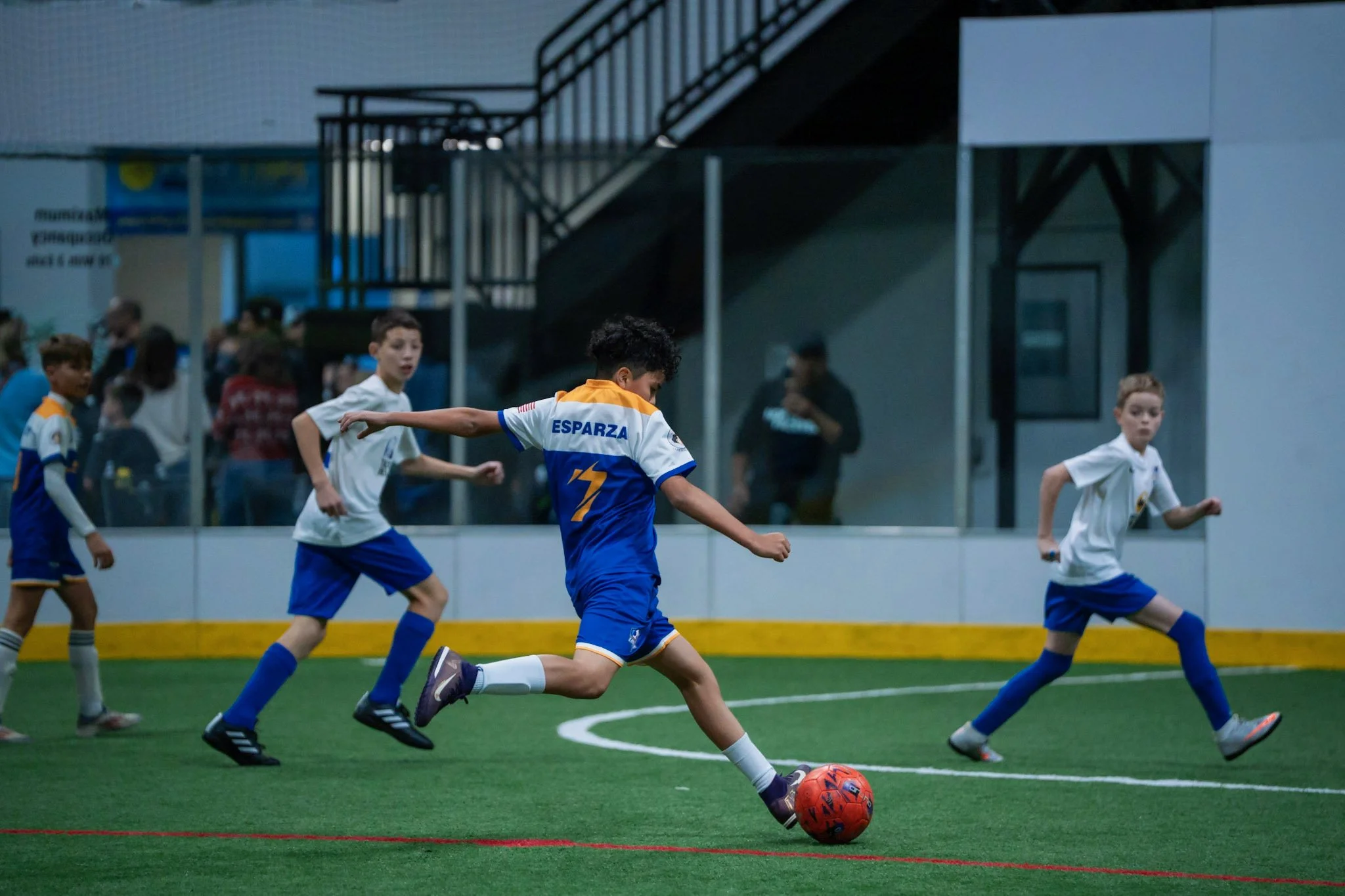 Young boys playing indoor soccer on artificial turf, with some watching from behind glass panels.