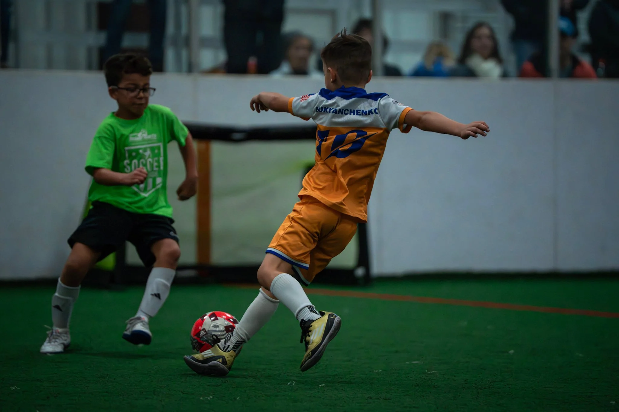 Two young boys playing indoor soccer, one in a green shirt and the other in an orange and blue uniform, competing for the ball.