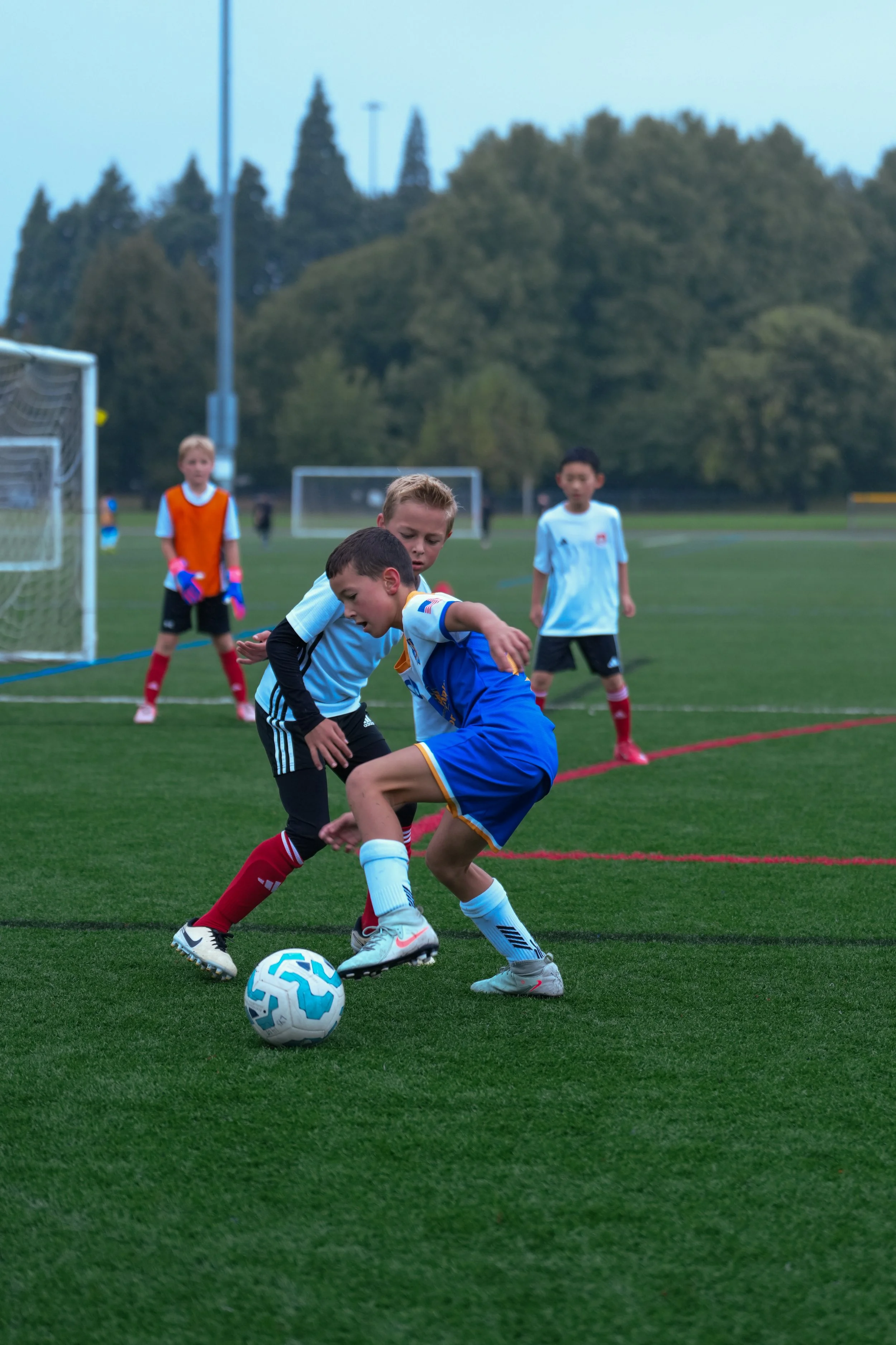 Children playing youth soccer on a field, with two boys fighting for control of the ball while others watch, in a park with trees in the background.